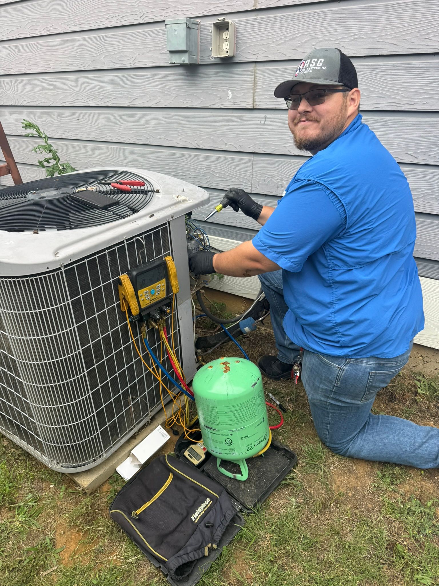 Technician kneeling beside outdoor AC unit, working on pipes and gauges next to a green refrigerant tank.