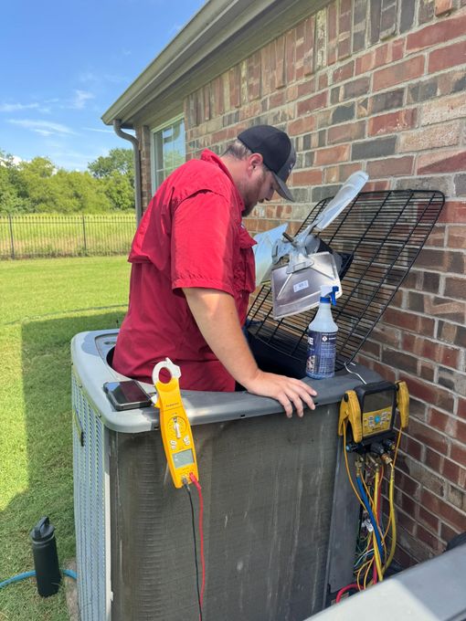 Technician in red shirt cleaning an outdoor HVAC unit beside a brick house