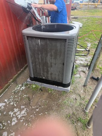 Outdoor AC unit being cleaned beside a red fence, with a worker in a blue shirt nearby