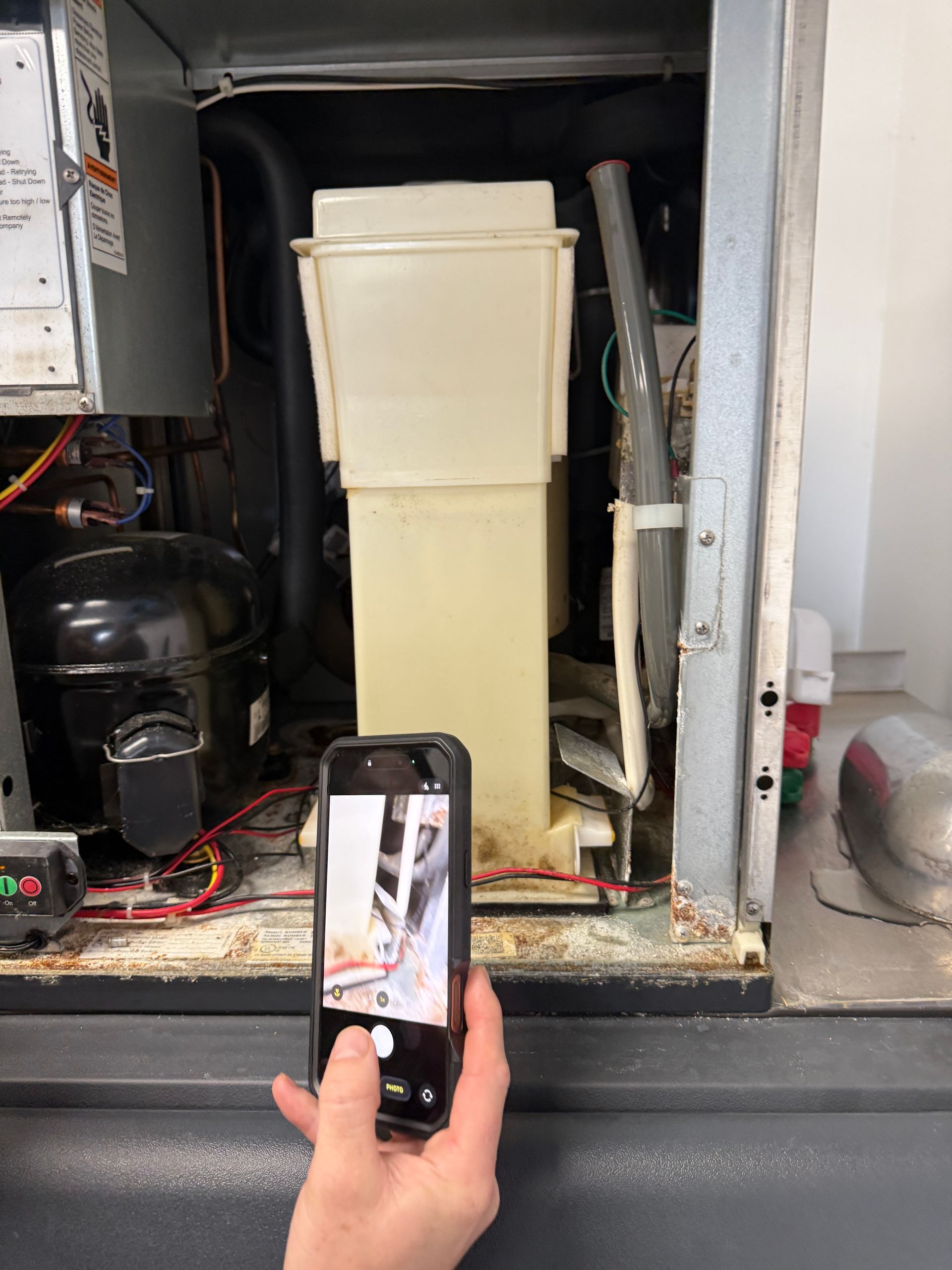 Hand holding a phone in front of an open HVAC unit with a beige filter or cabinet inside