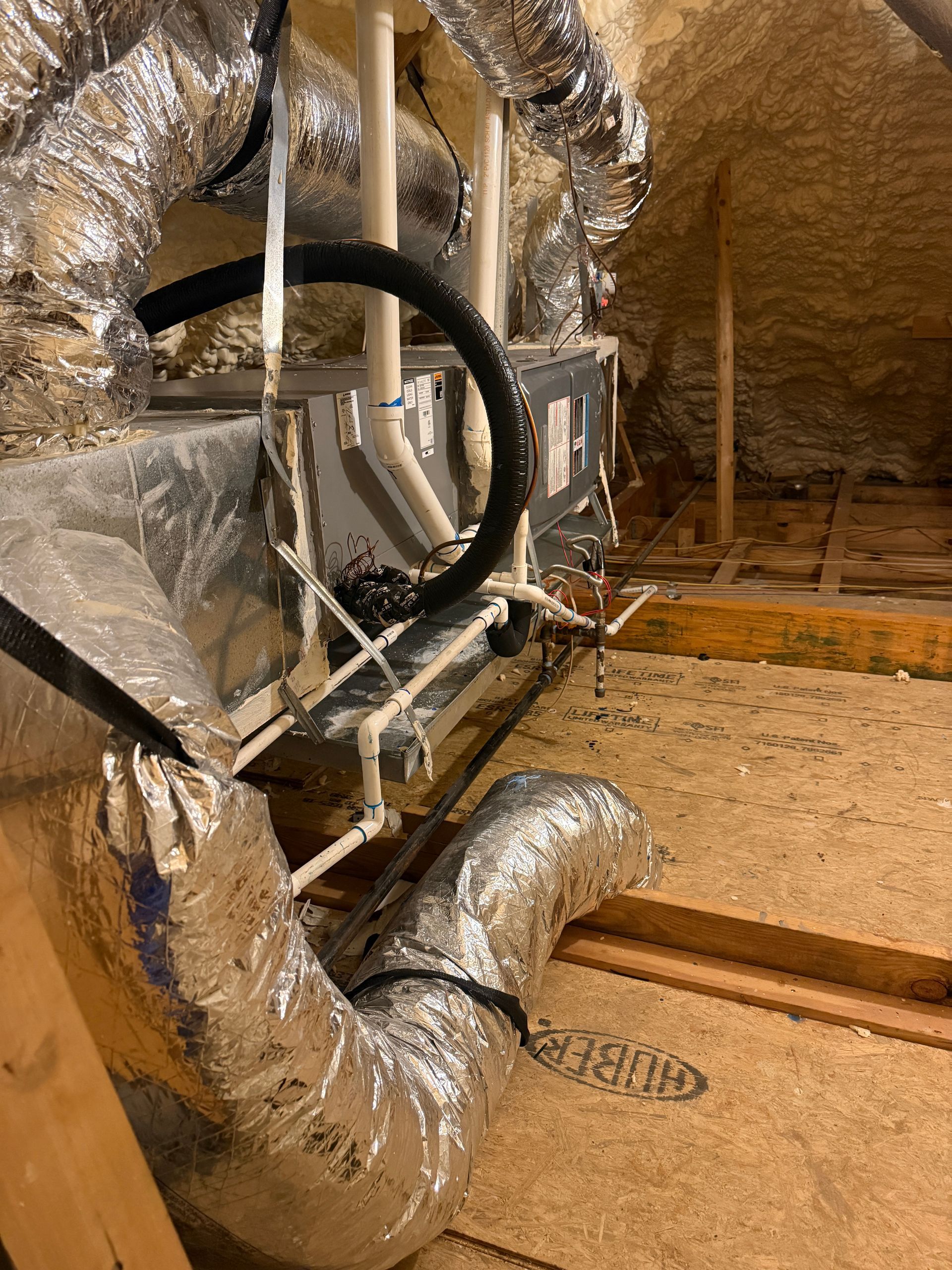 Attic HVAC ductwork with insulated silver ducts and black hoses near exposed wooden rafters.
