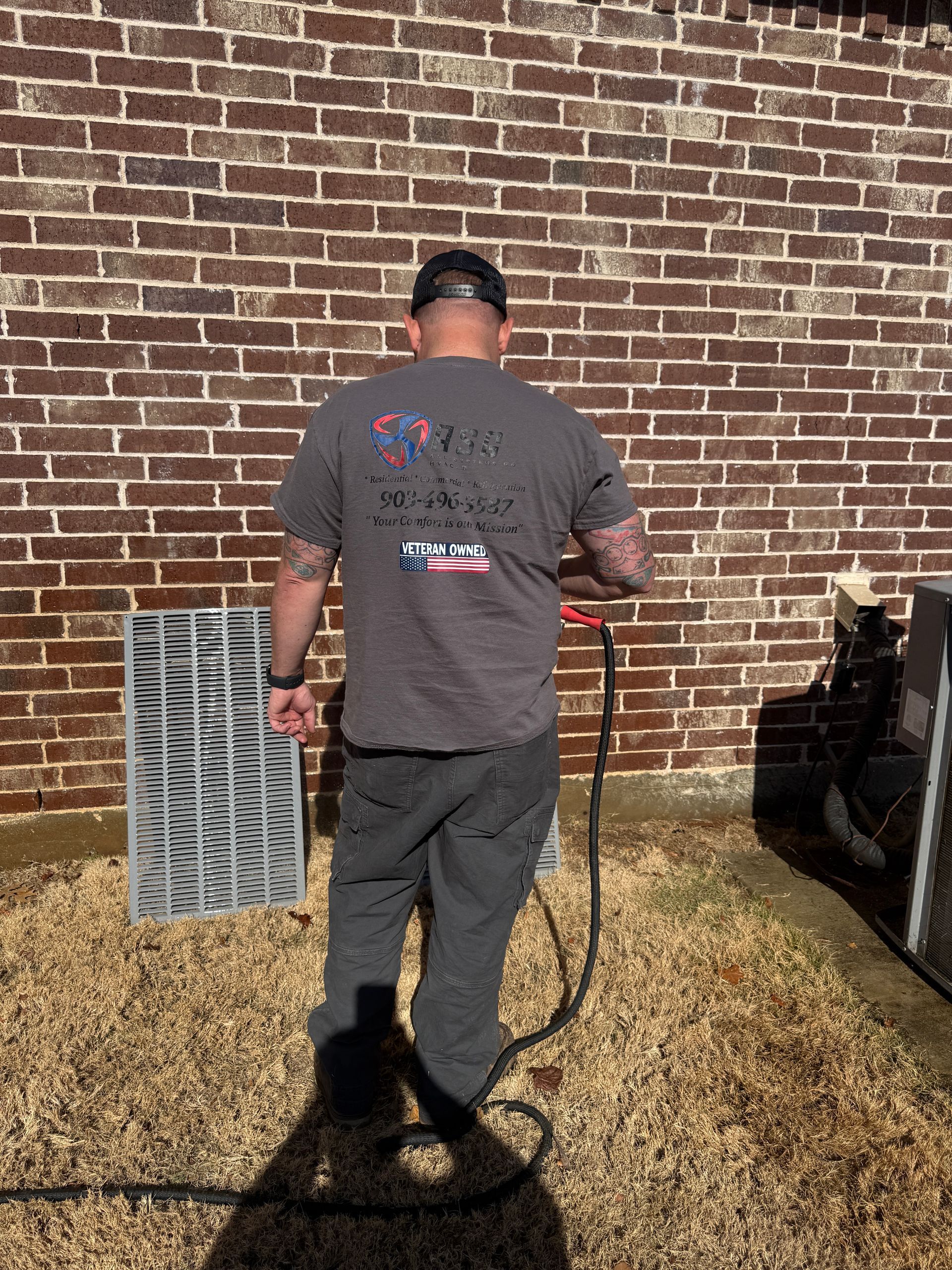 Person in a cap standing by a brick wall, holding a shovel in a yard with dry grass.