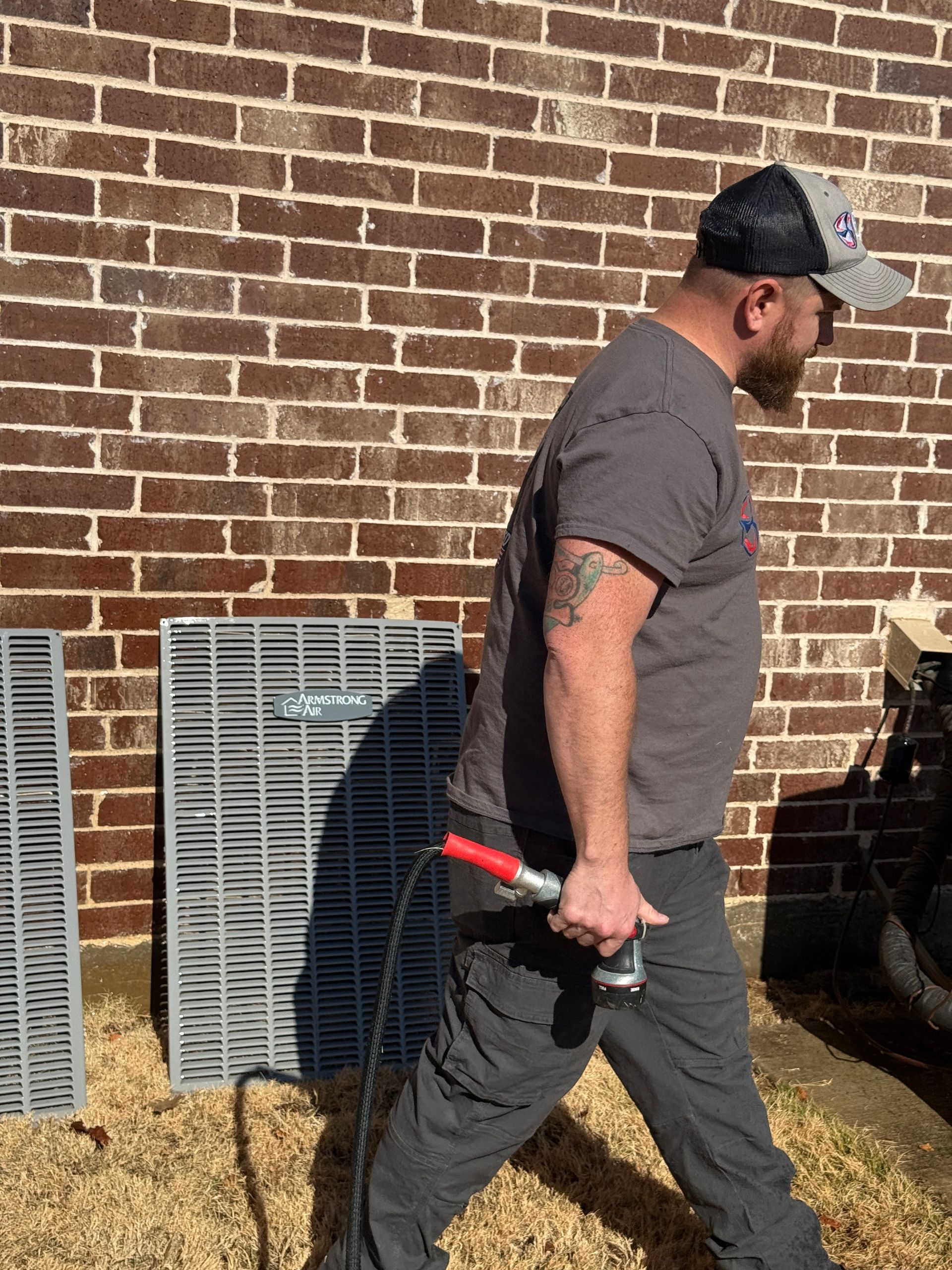 Man in gray clothes and cap carrying a red-handled tool beside a brick wall and HVAC units