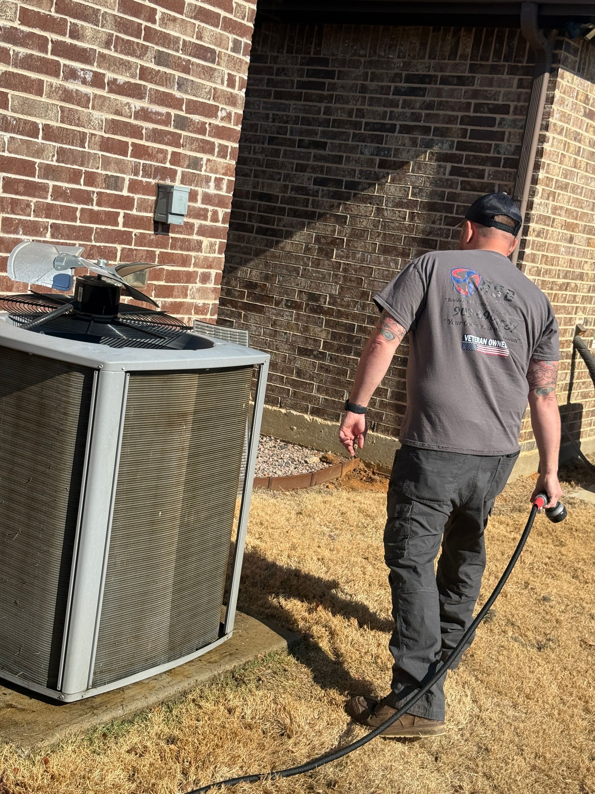 Technician walking beside an outdoor HVAC unit near a brick wall, holding a hose or tool.
