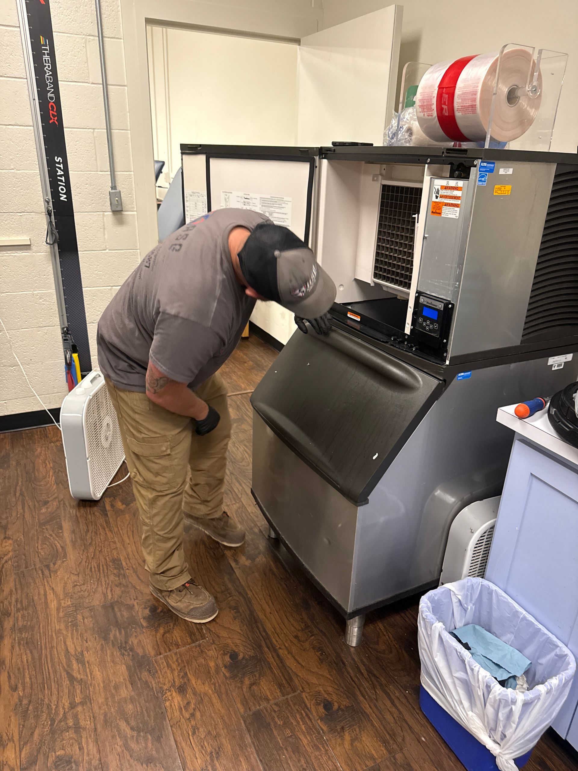 Person inspecting a large ice machine in a utility room, with trash bins and supplies nearby.