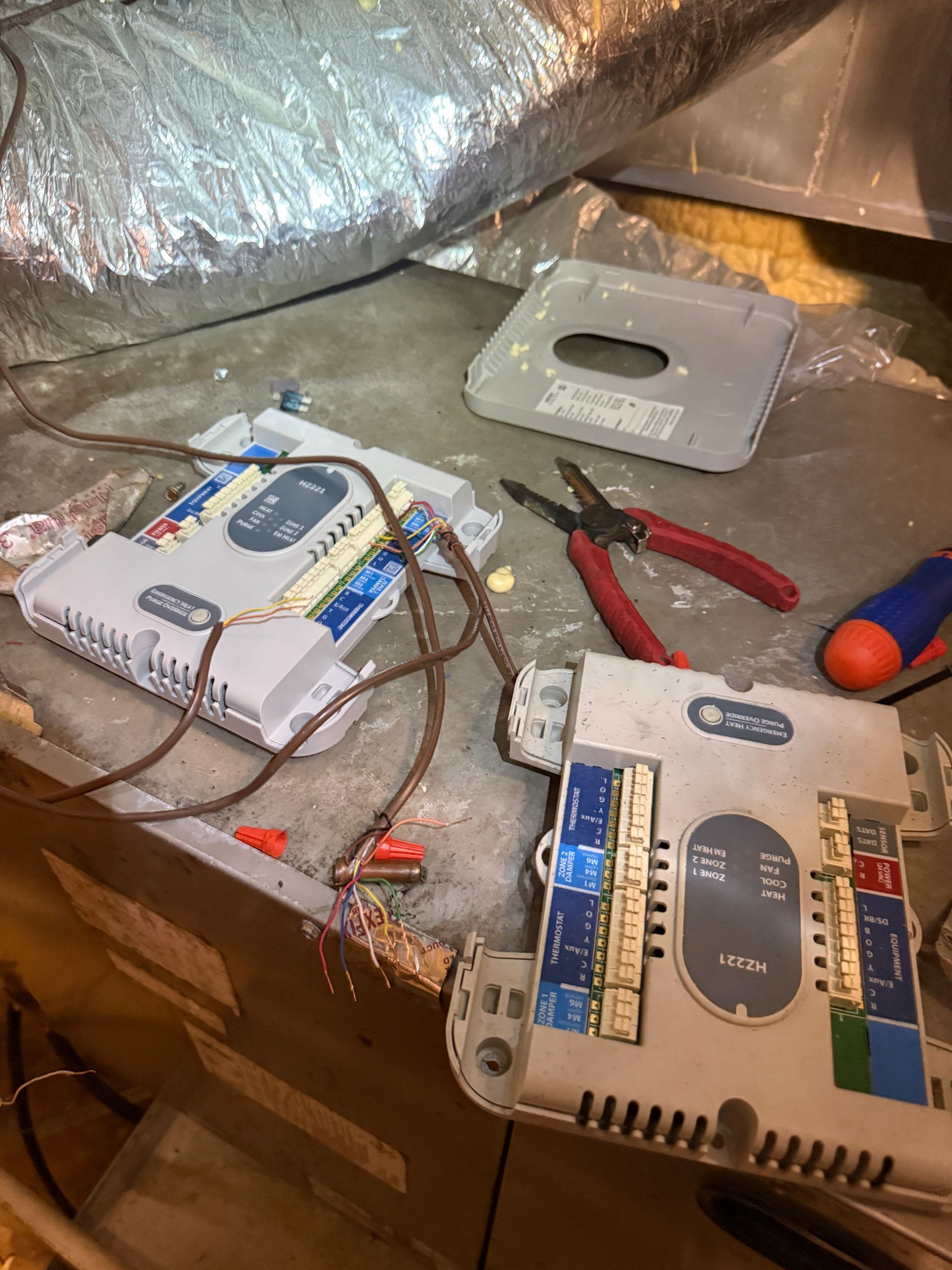 Two metal electrical boxes with wires and tools on a dusty floor beside silver ducting.