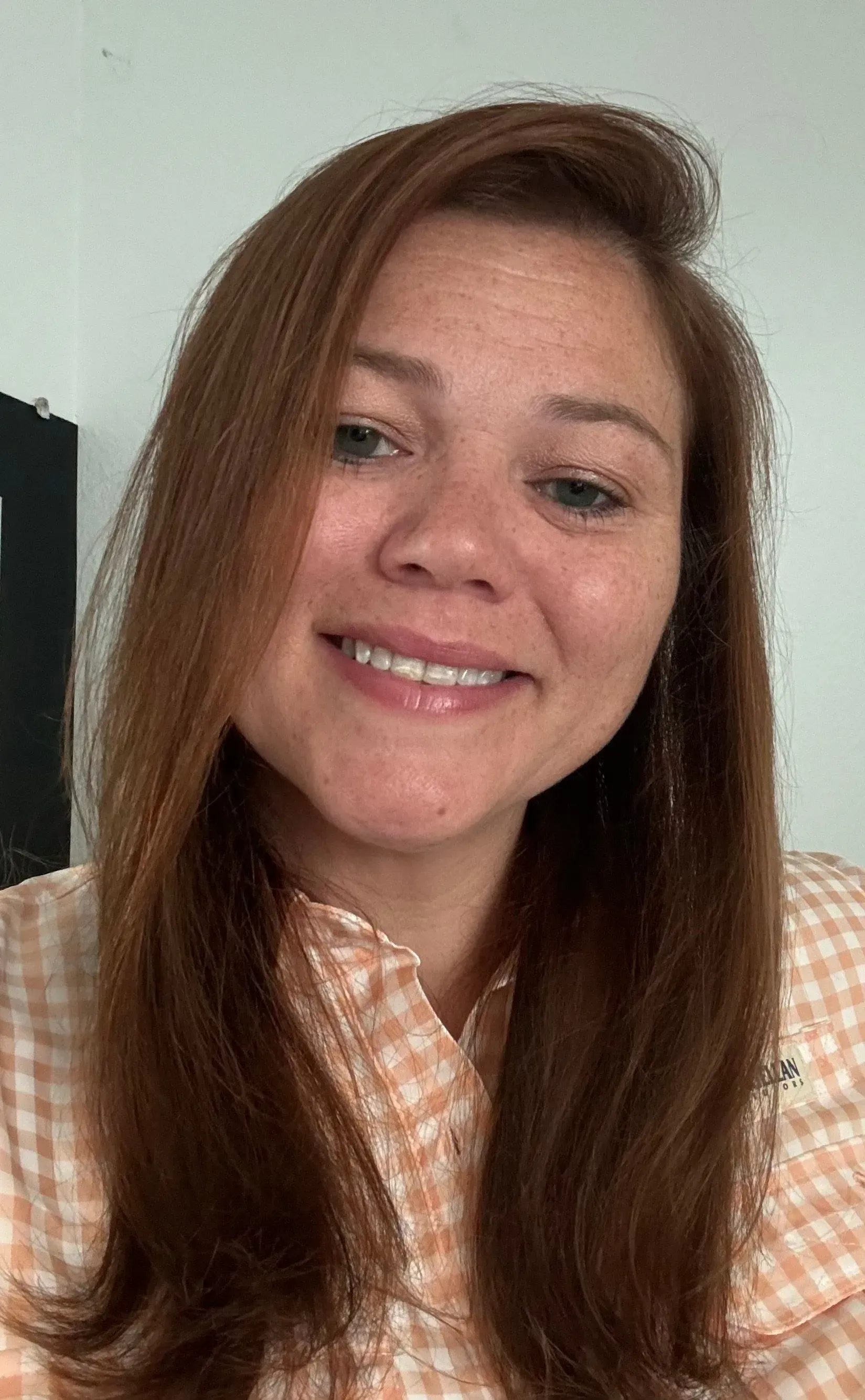 Smiling person with long brown hair wearing a peach plaid shirt against a light background
