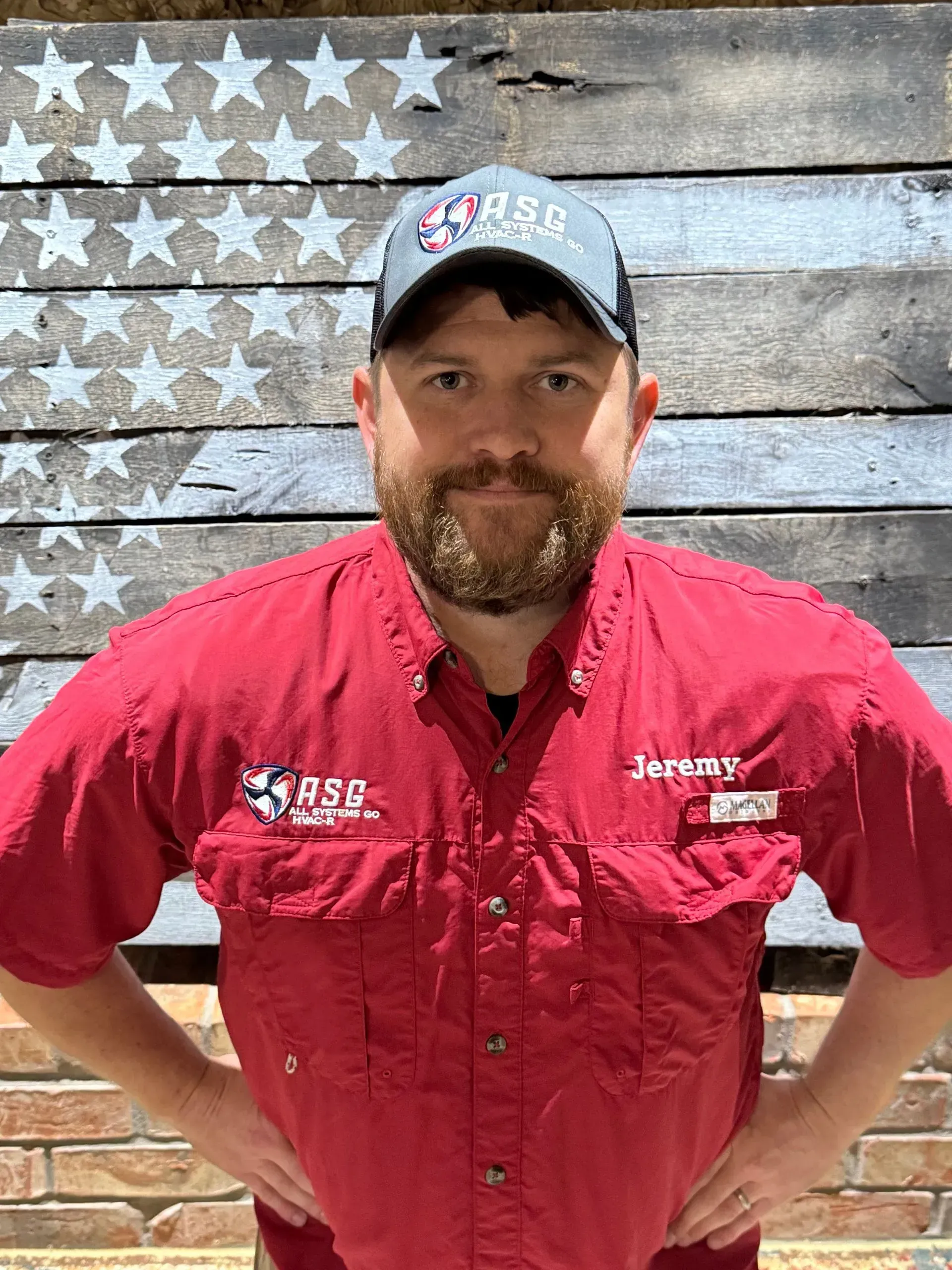 Man in red work shirt and cap standing before a weathered American flag backdrop.