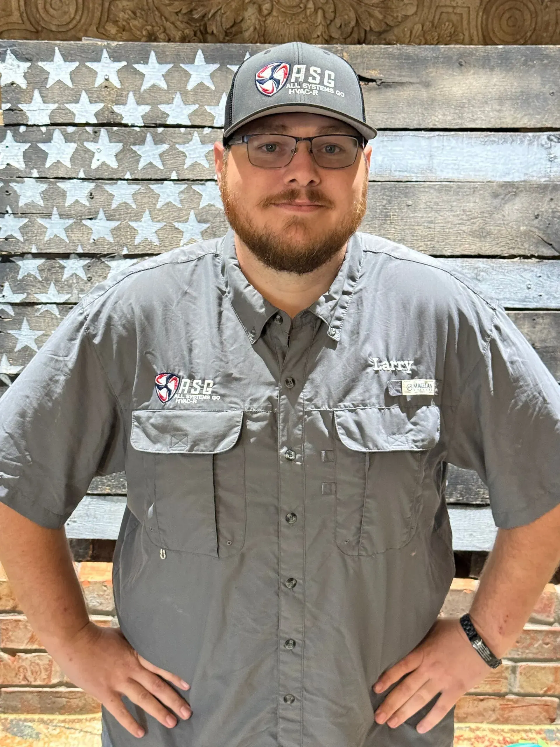 Man in gray shirt and cap, hands on hips, standing before a weathered American flag backdrop.