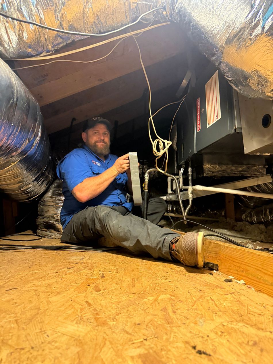 Worker in blue shirt kneeling under a crawlspace, using tools beside insulation and exposed beams