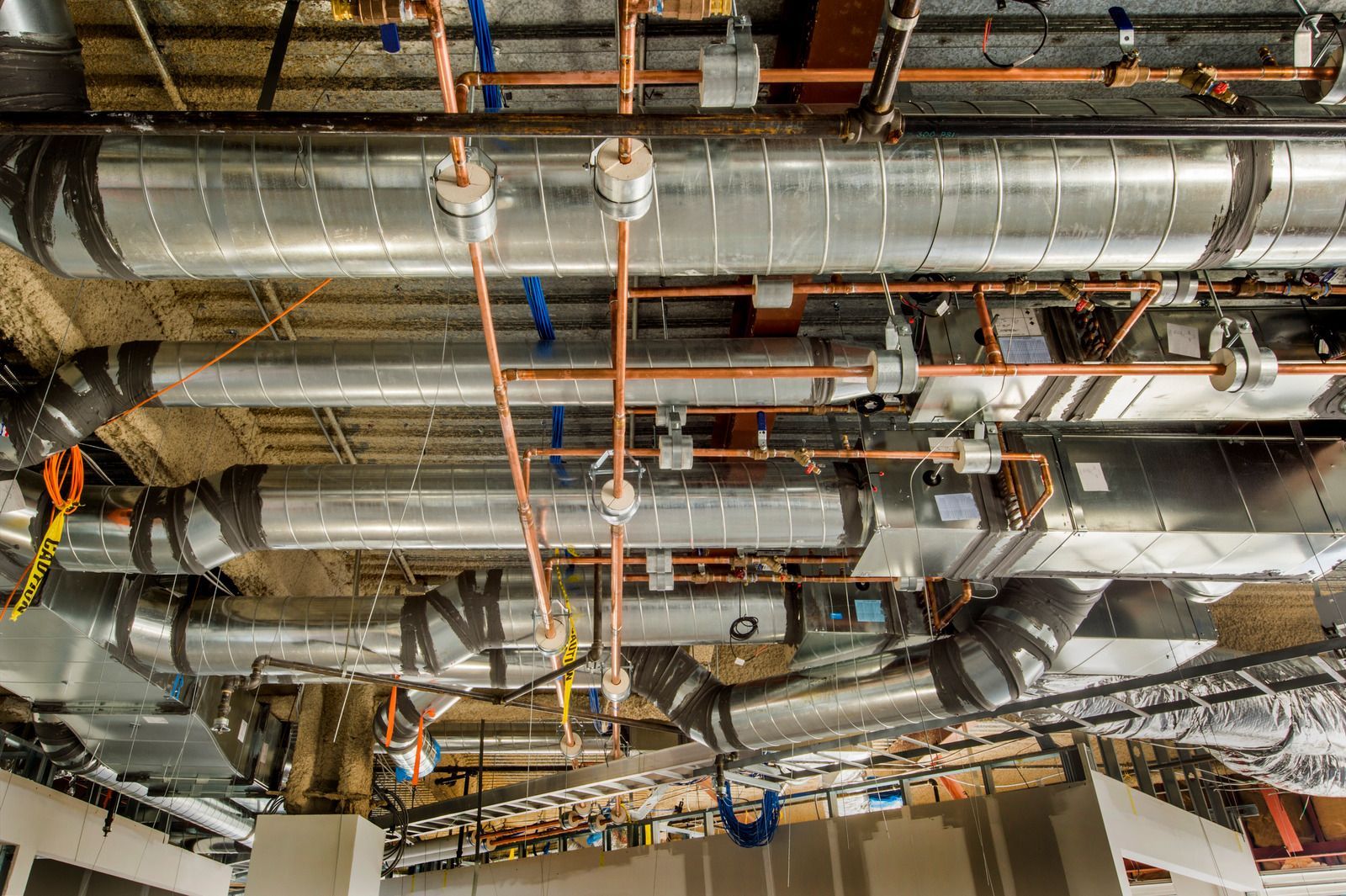 Industrial ceiling with exposed silver ductwork, pipes, and orange electrical conduits in a construction area