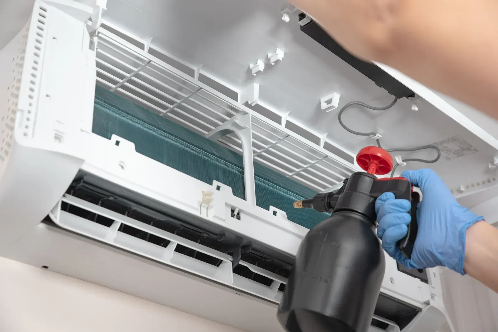 Technician spraying cleaner into a wall-mounted air conditioner’s open interior with a red nozzle bottle.
