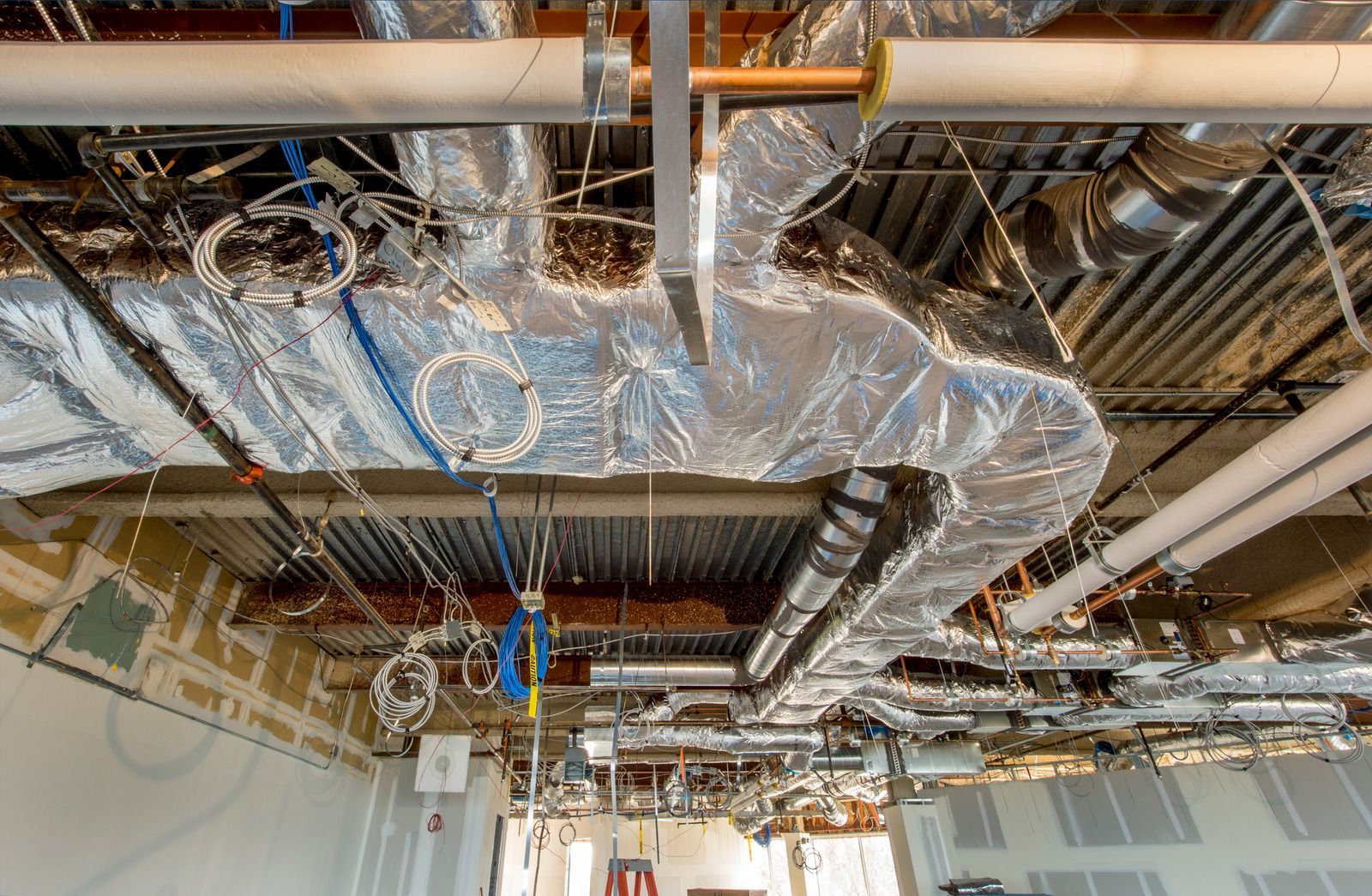 Exposed HVAC ducts and pipes in an unfinished ceiling with reflective insulation and hanging electrical wiring