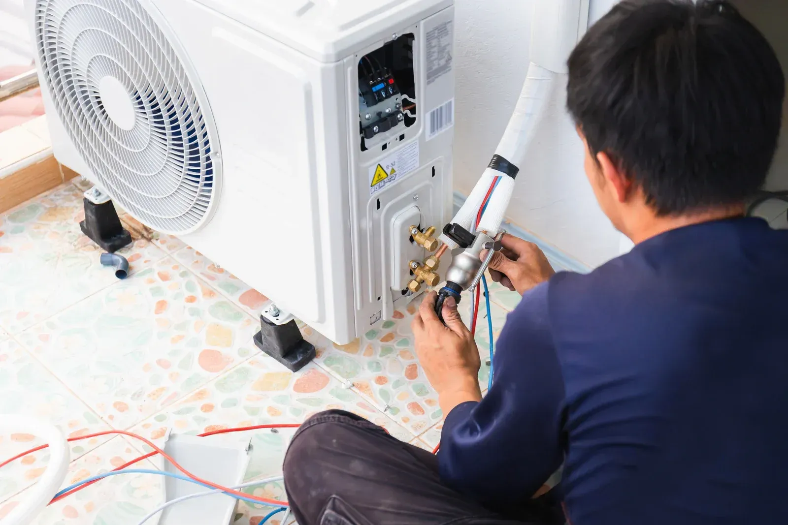 Technician repairing a white outdoor air conditioner unit with tools on a tiled floor