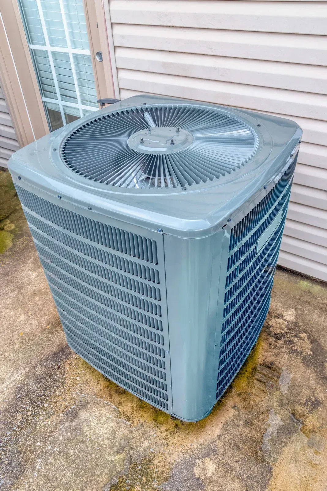 Outdoor air conditioner unit beside a house, gray metal with top fan and side vents.