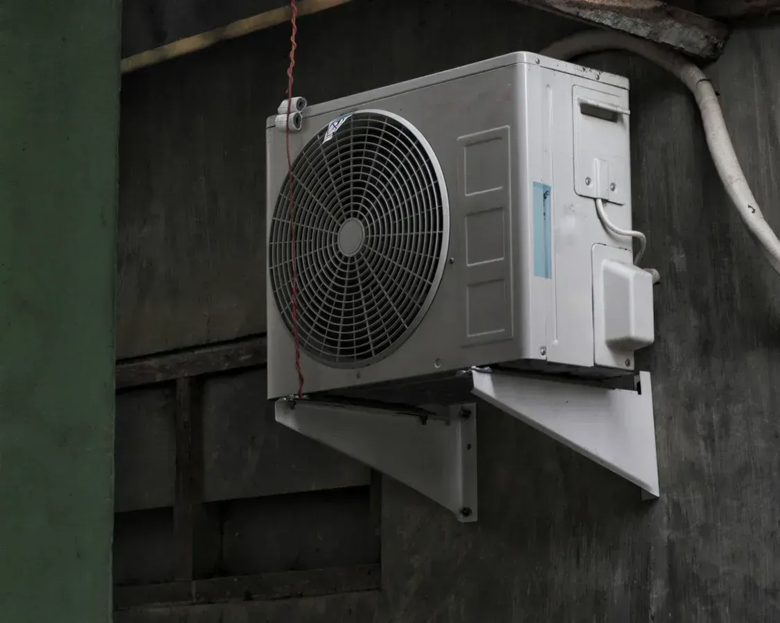 Outdoor wall-mounted air conditioner unit with a large fan on a green industrial wall.