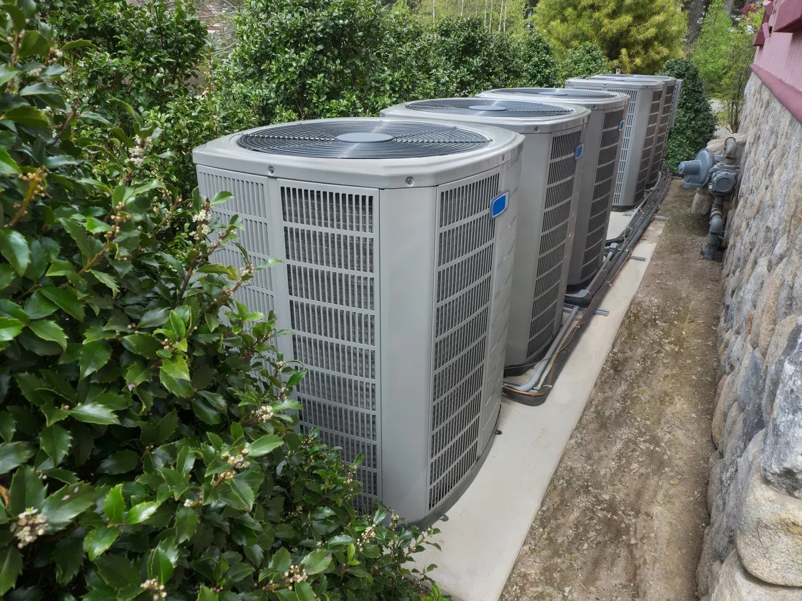 Row of outdoor air conditioning units beside a stone wall and greenery