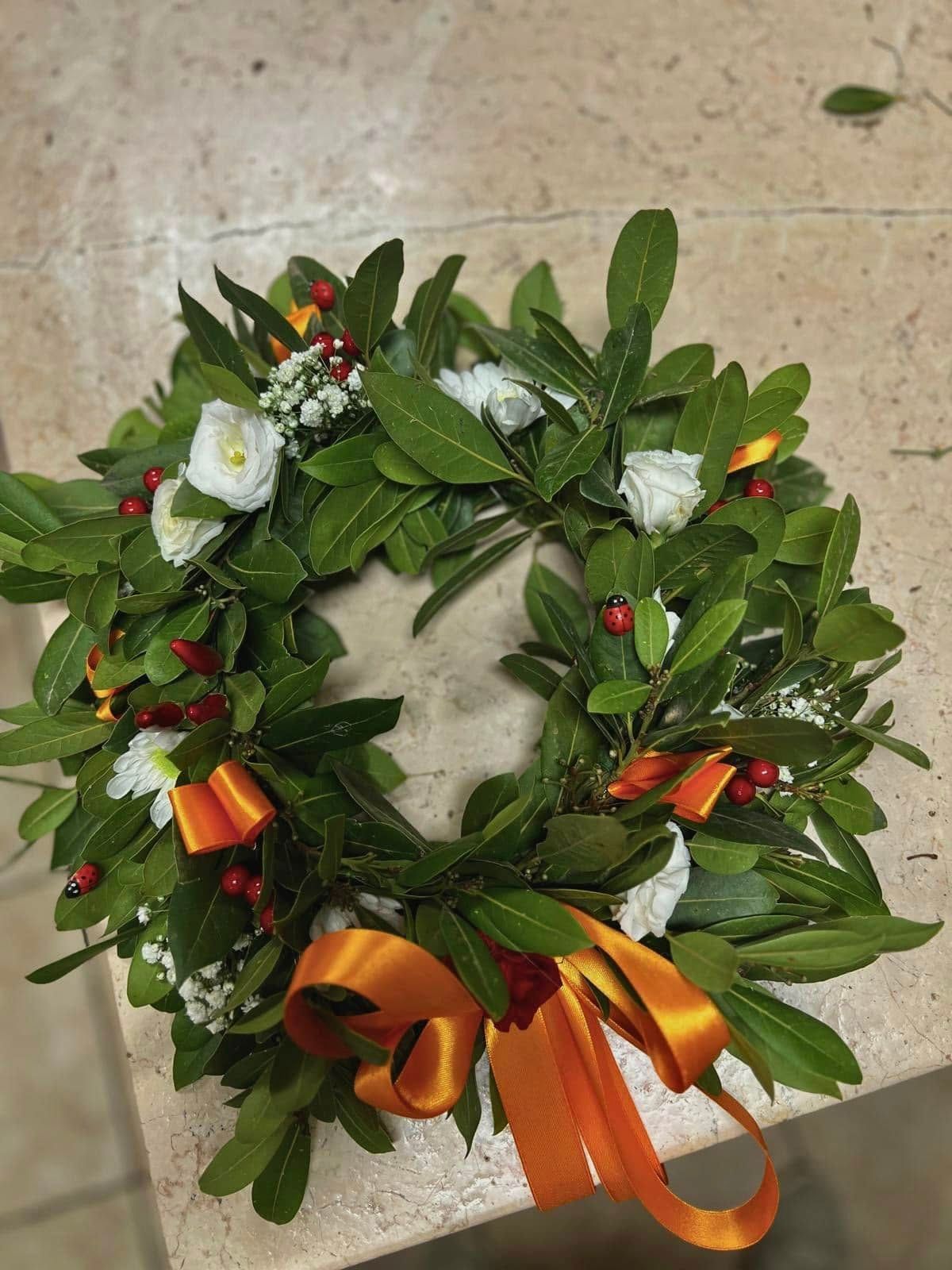 Ghirlanda di foglie verdi, fiori bianchi, nastro arancione e bacche su una superficie di pietra.