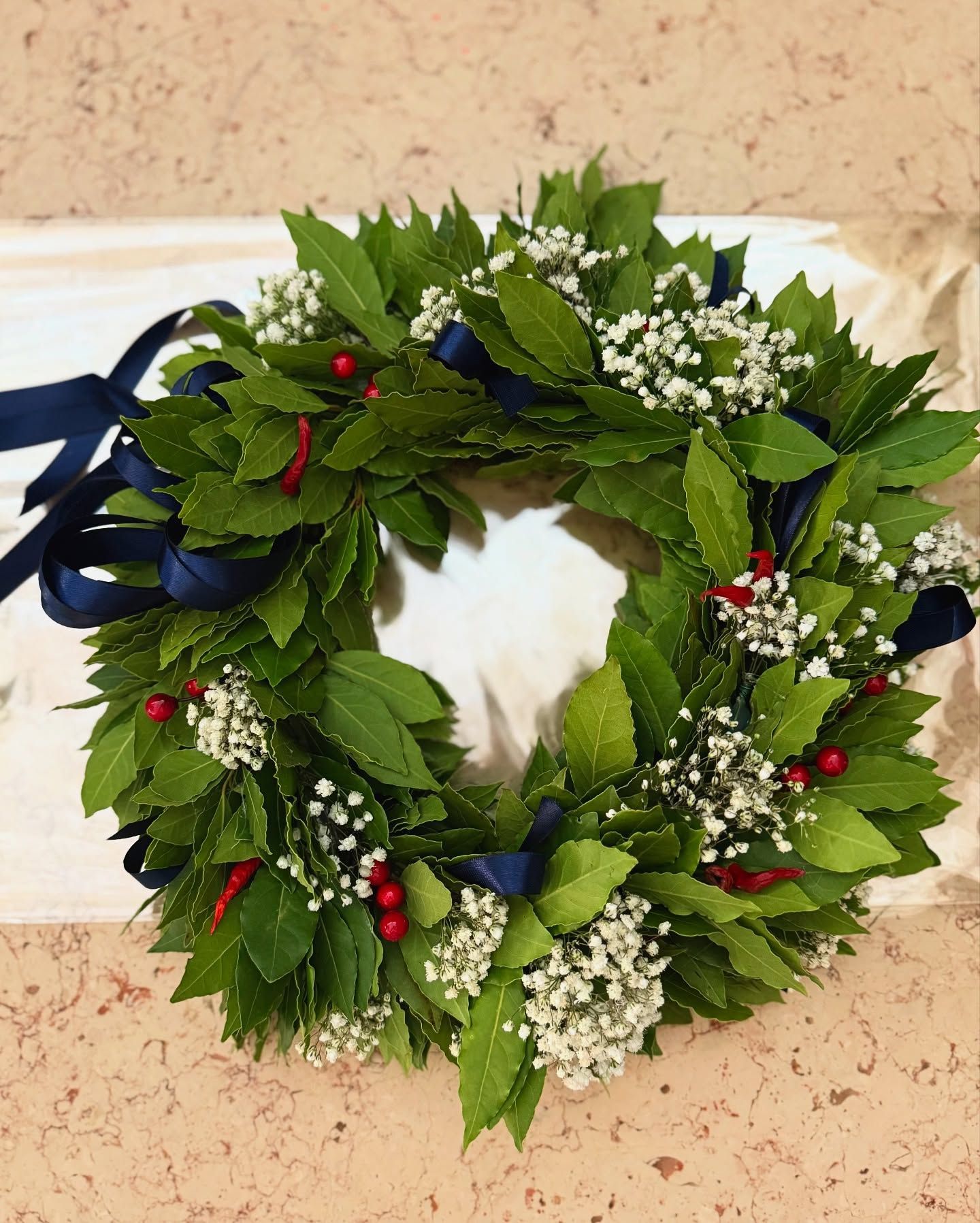 Una corona d'alloro con foglie verdi, piccoli fiori bianchi, bacche rosse e nastri blu.