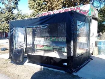 Carpa plegable de malla negra con laterales de plástico transparente en una acera soleada frente a una tienda.