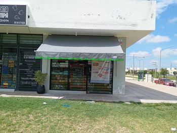 Pequeño local comercial con toldo verde, puertas de cristal y acera junto a un arcén cubierto de césped.