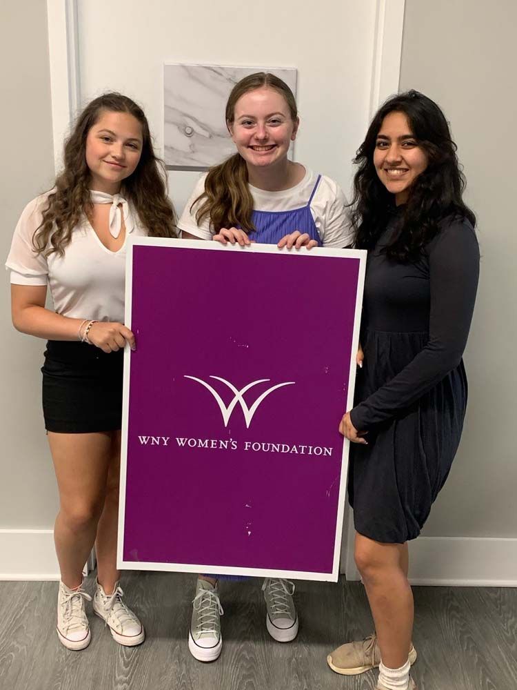 Three women are standing next to each other holding a purple sign.