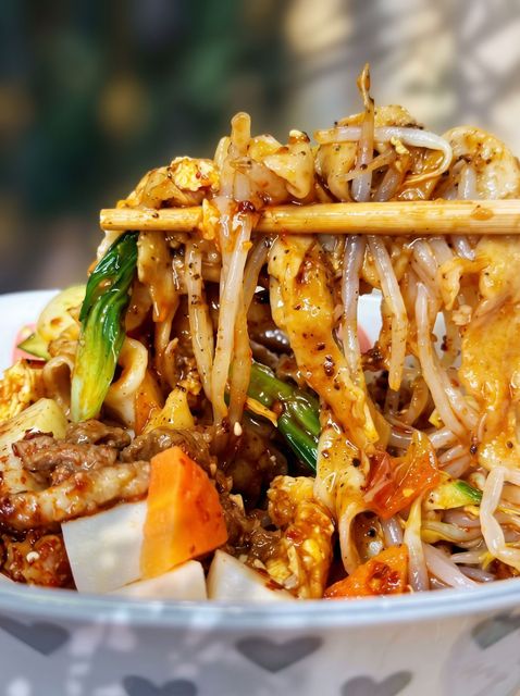 Close-up of stir-fried noodles with vegetables, held by chopsticks over a bowl
