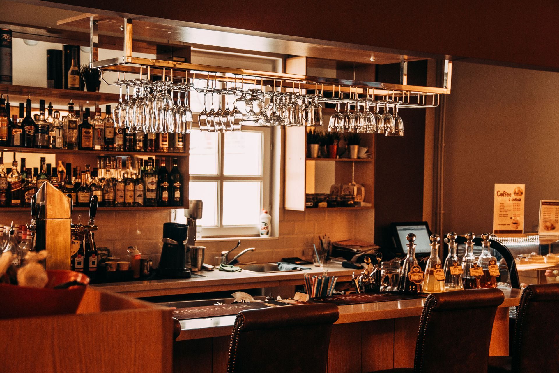 Warmly lit bar interior with shelves of bottles, hanging glasses, and a window behind the counter.