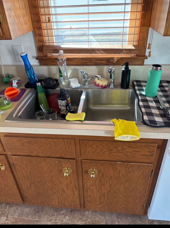 Kitchen sink and counter cluttered with dishes, bottles, and cleaning items beneath a window with blinds.