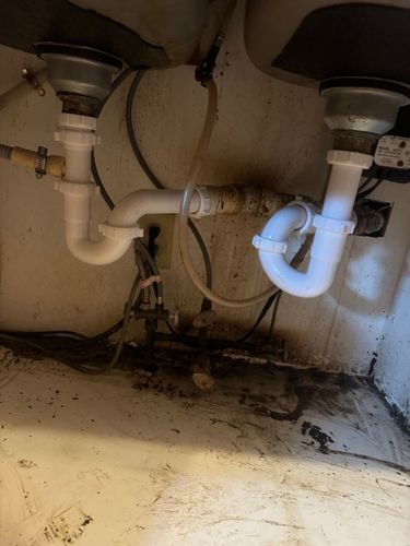 Under-sink plumbing with exposed pipes, a P-trap, and dirty cabinet floor with stains and debris