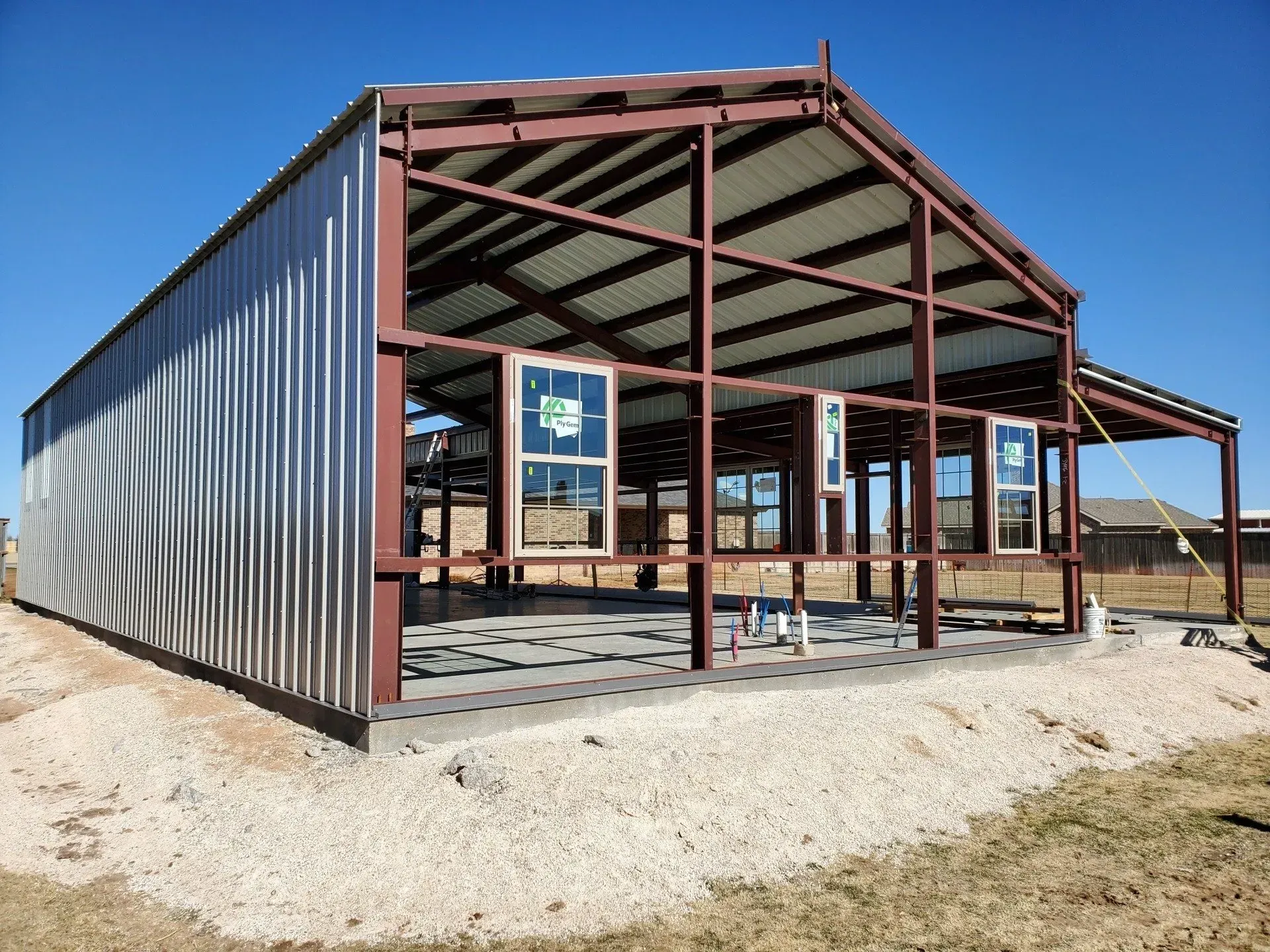 Steel-framed building under construction with corrugated metal siding on a gravel lot under a clear blue sky