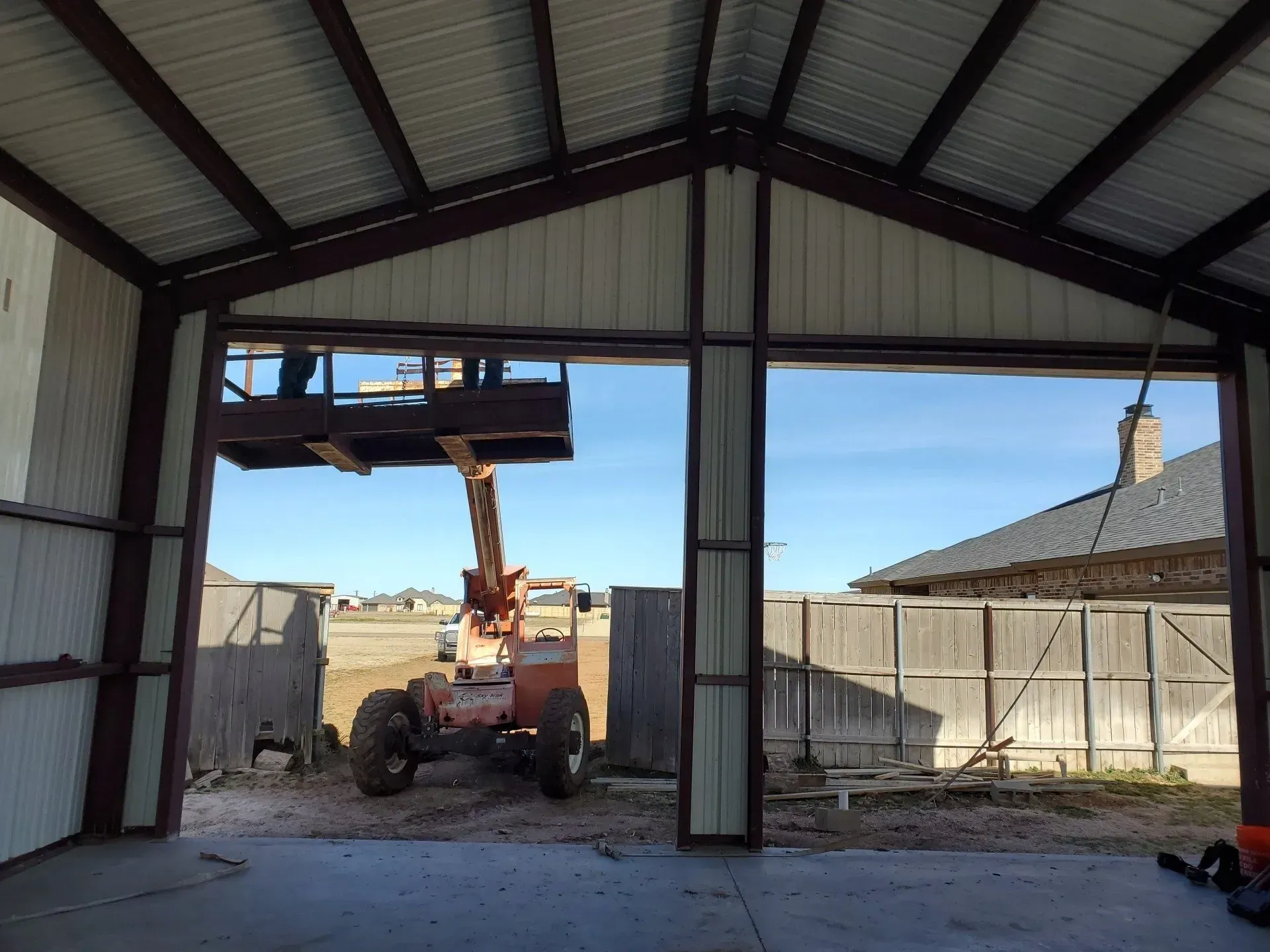 Forklift lifting materials inside an open metal shed, with a fenced yard visible outside.