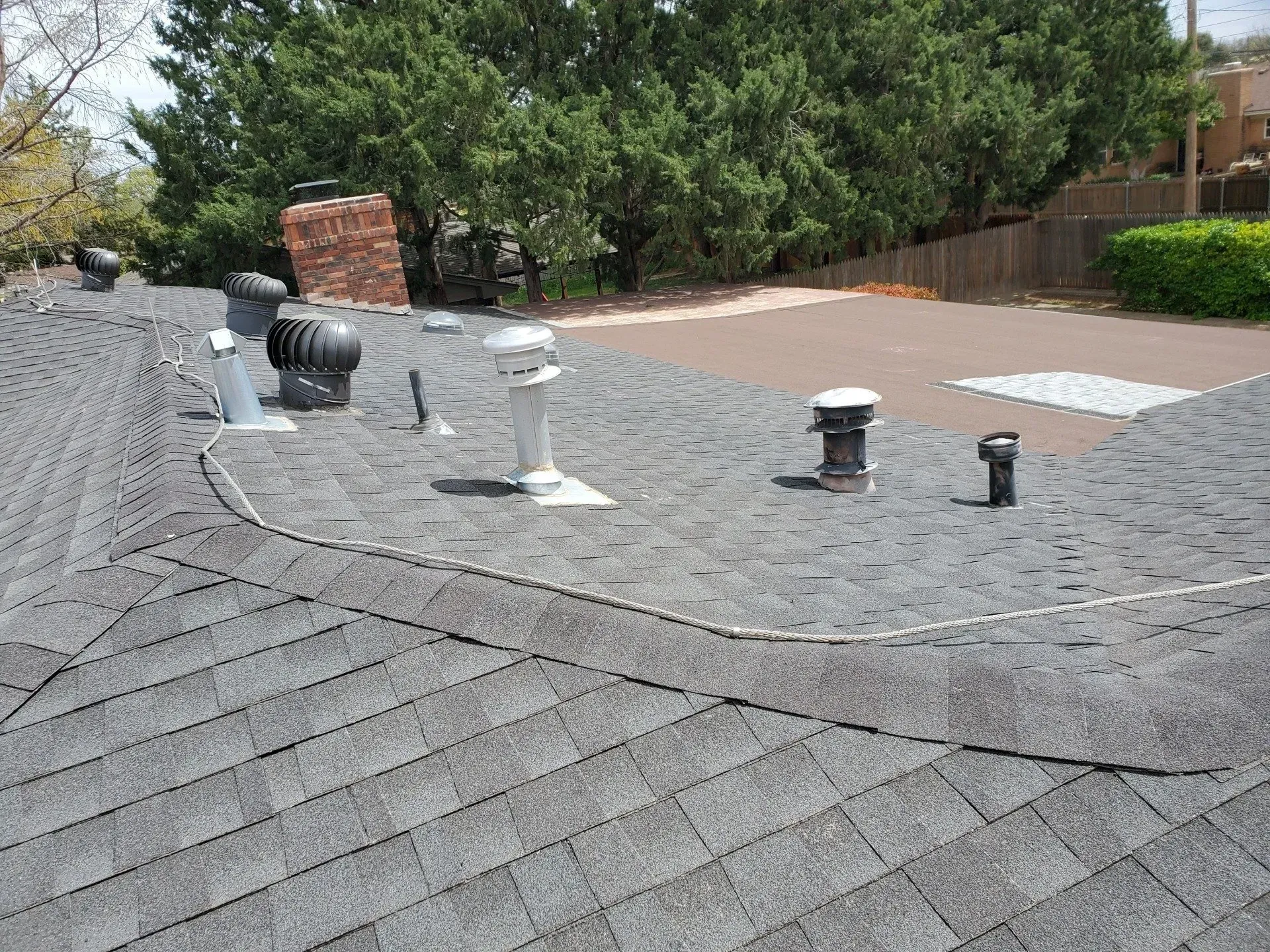 Roof with multiple vents and a chimney, viewed from above with trees and a driveway in the background