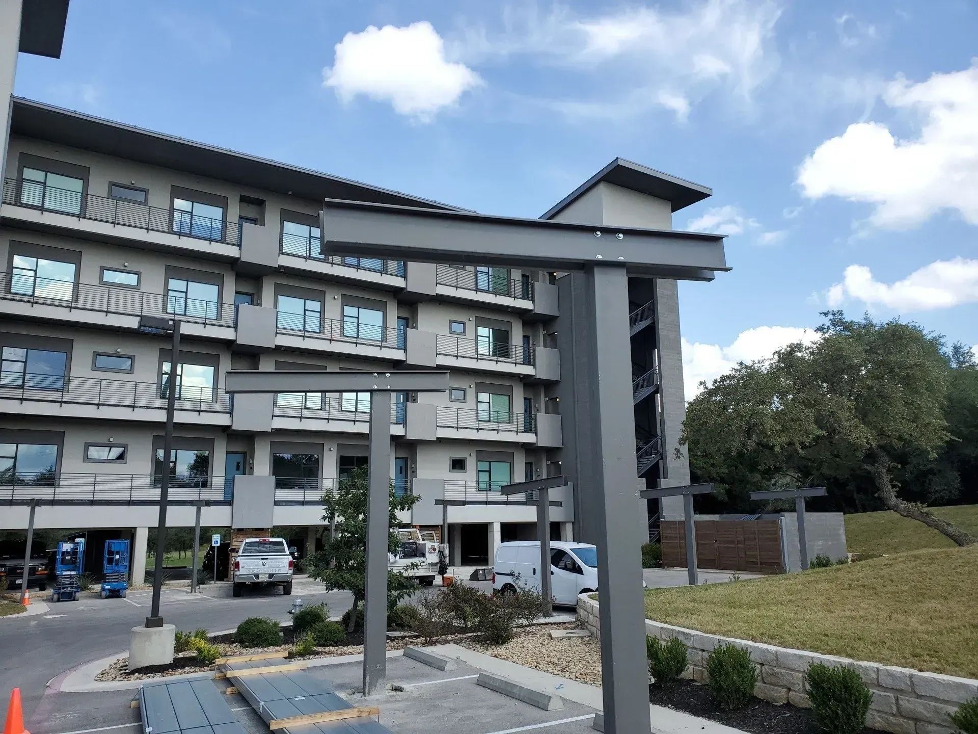 Modern apartment building with balconies and parked cars, viewed from a landscaped courtyard under a blue sky