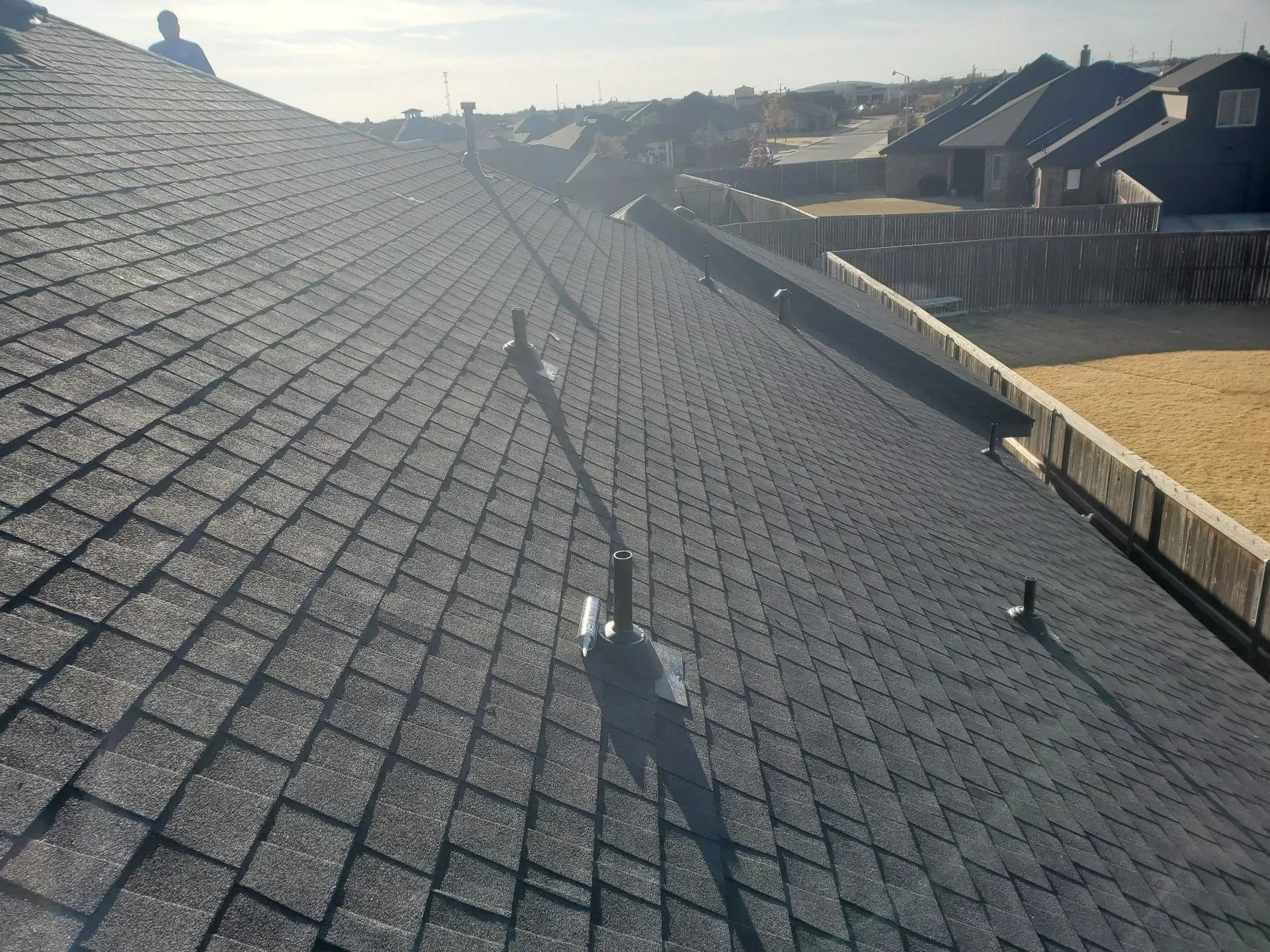 Roof covered in dark gray shingles with two vent pipes, viewed from above in bright sunlight