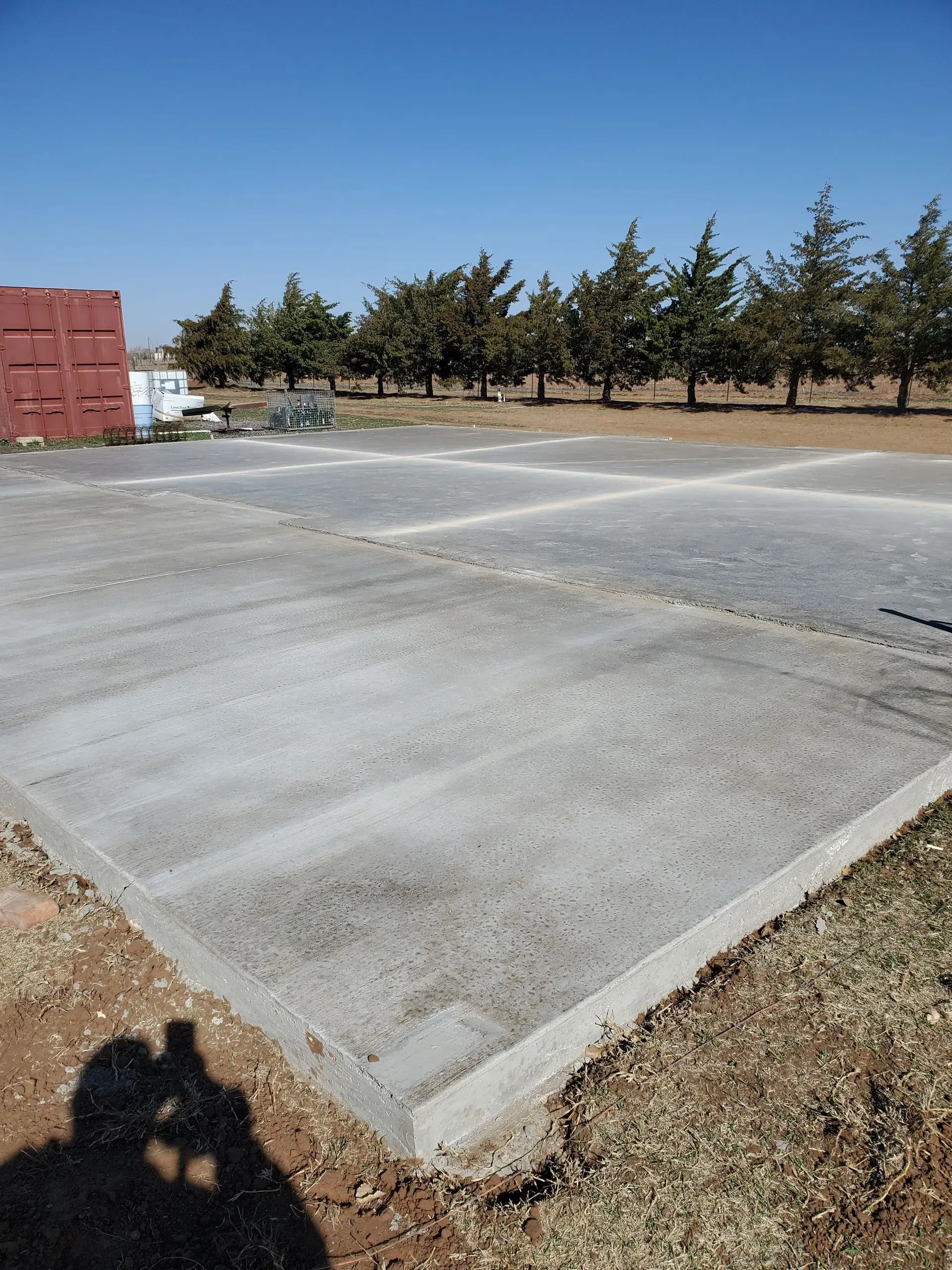 Empty concrete lot with a red container and trees under a clear blue sky.