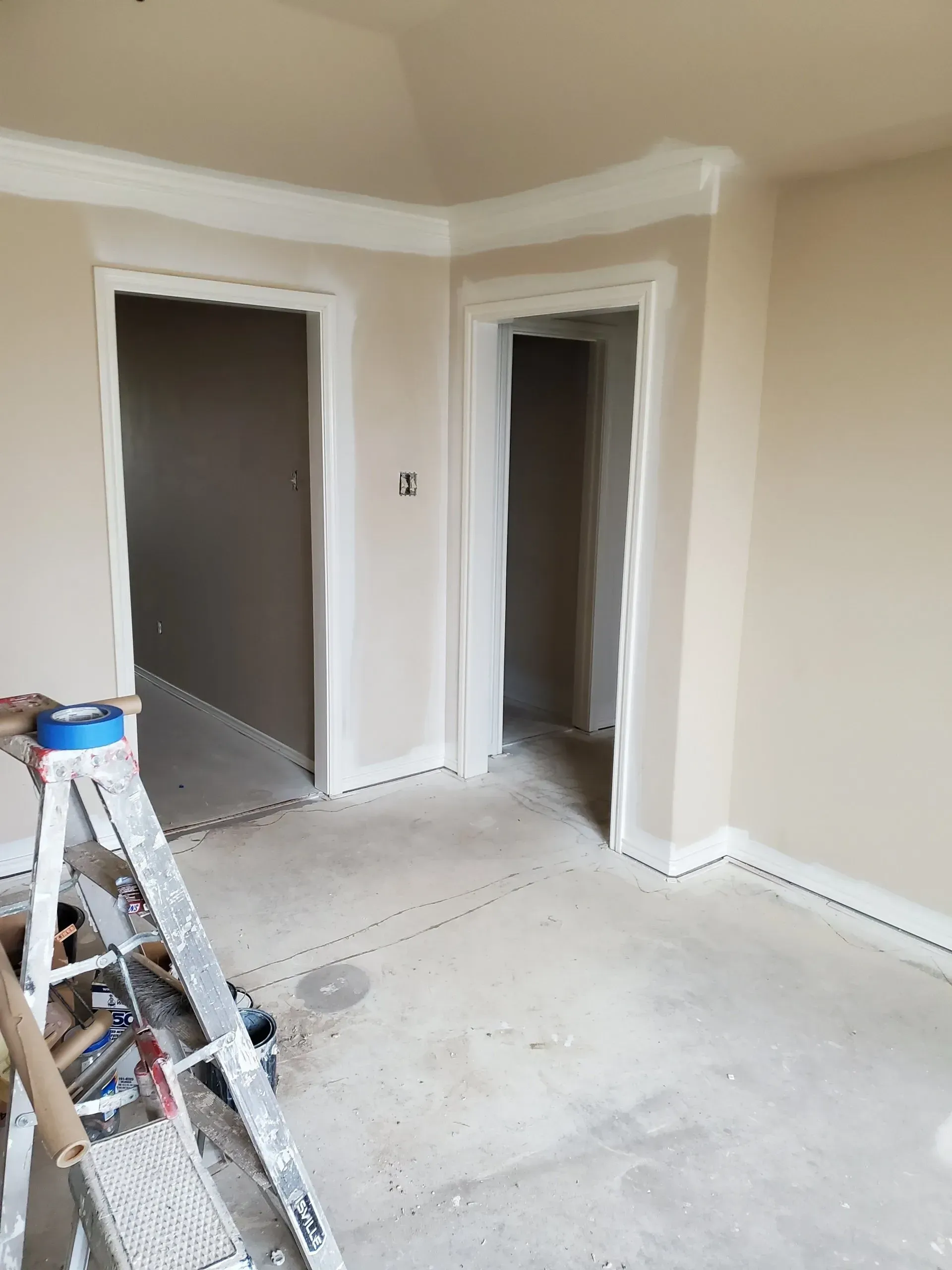 Unfinished room with beige walls, open doorways, and a ladder on a dusty floor near a ceiling edge.