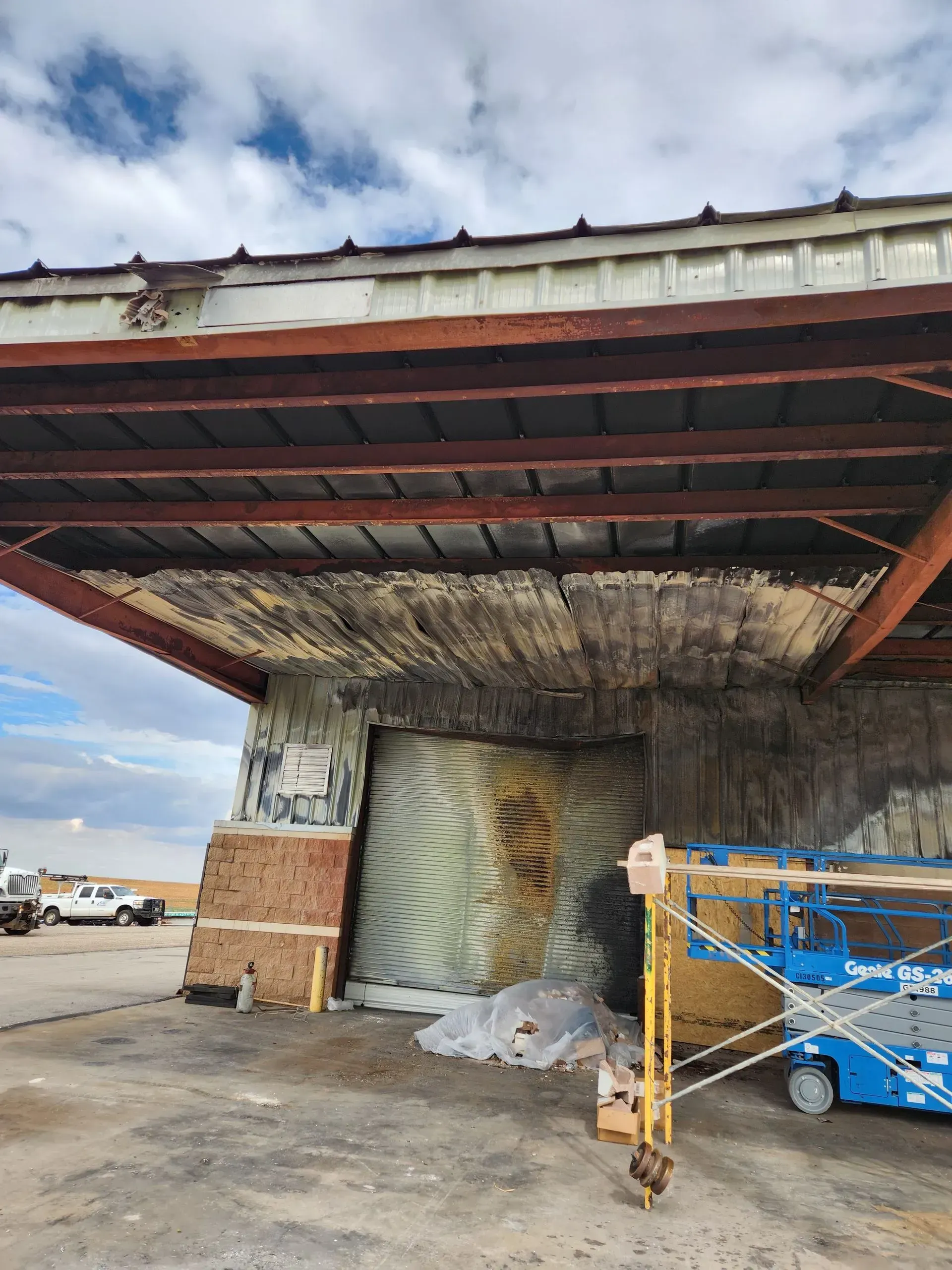 Dilapidated industrial awning with peeling concrete, rusted metal, and a blue cart on a concrete lot