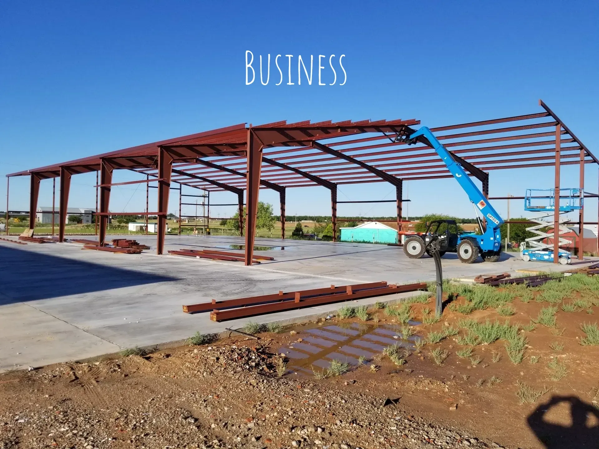 Steel building frame under construction with a blue lift on a concrete slab in a sunny lot
