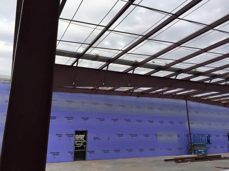 Empty indoor construction space with blue wall sheathing, steel beams, and a skylit roof