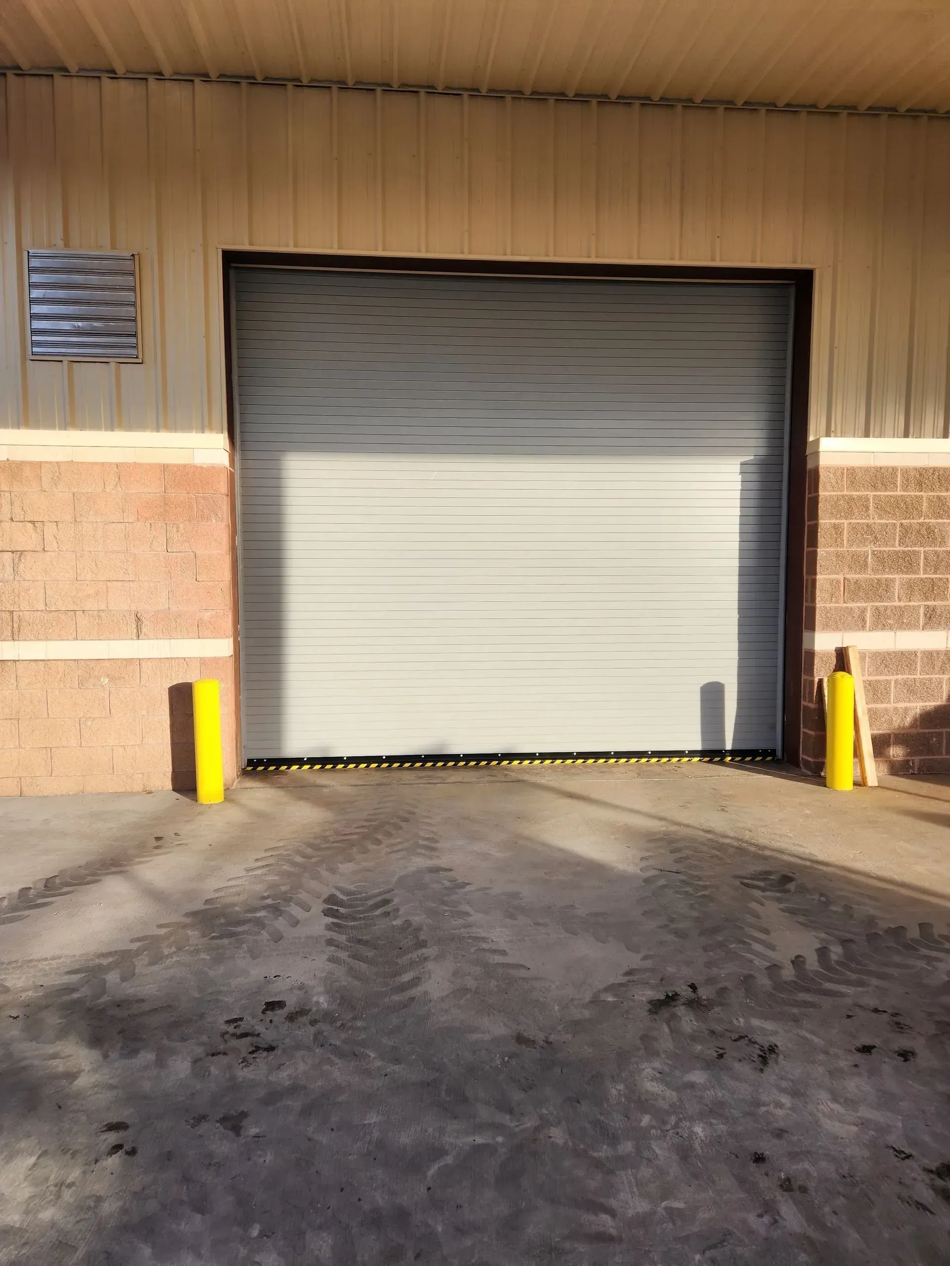 Closed gray roll-up garage door at a warehouse loading bay with yellow bollards and a concrete driveway