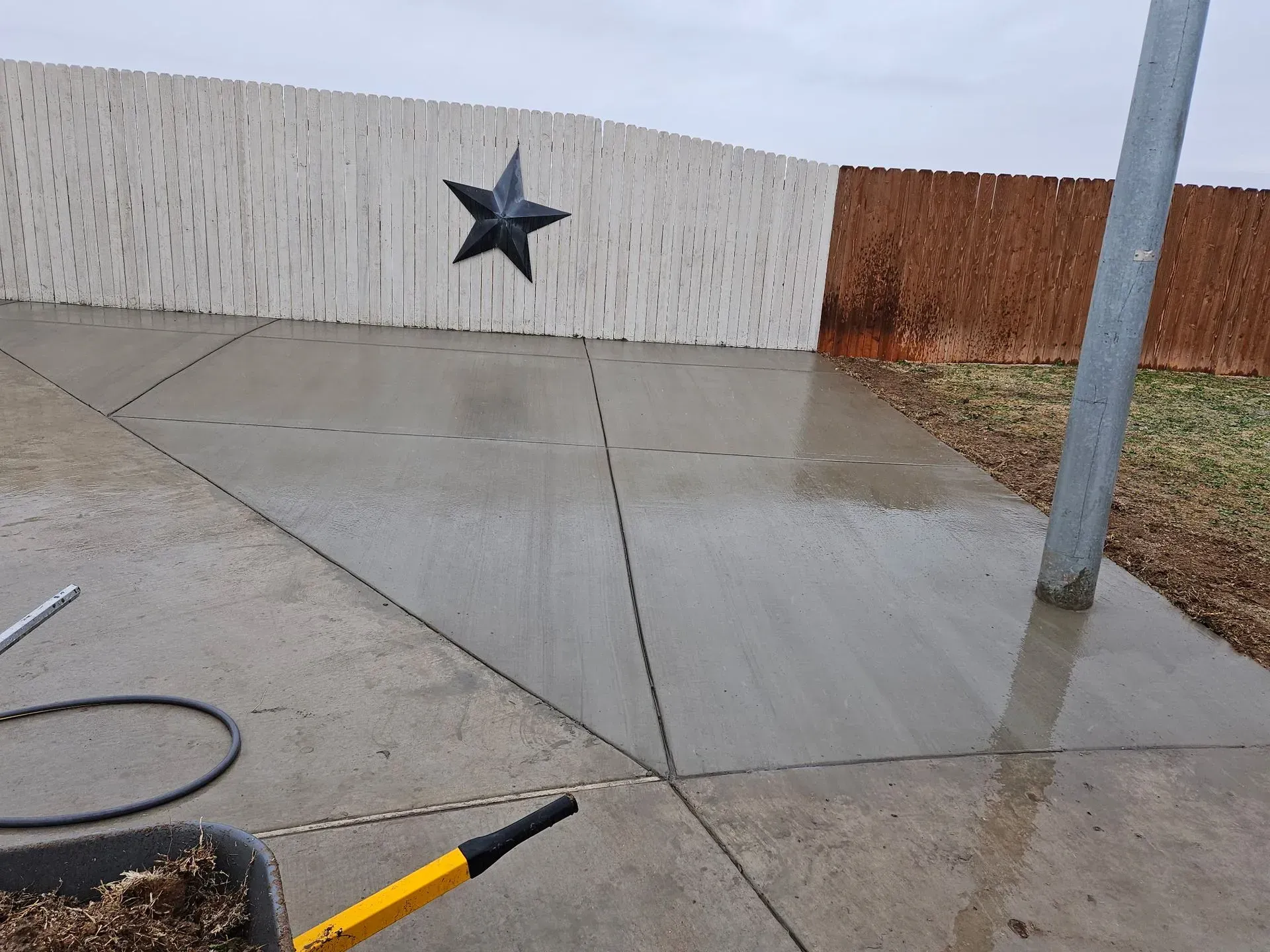 Wet concrete walkway beside a white wall with a black star, rusted fence, and pole