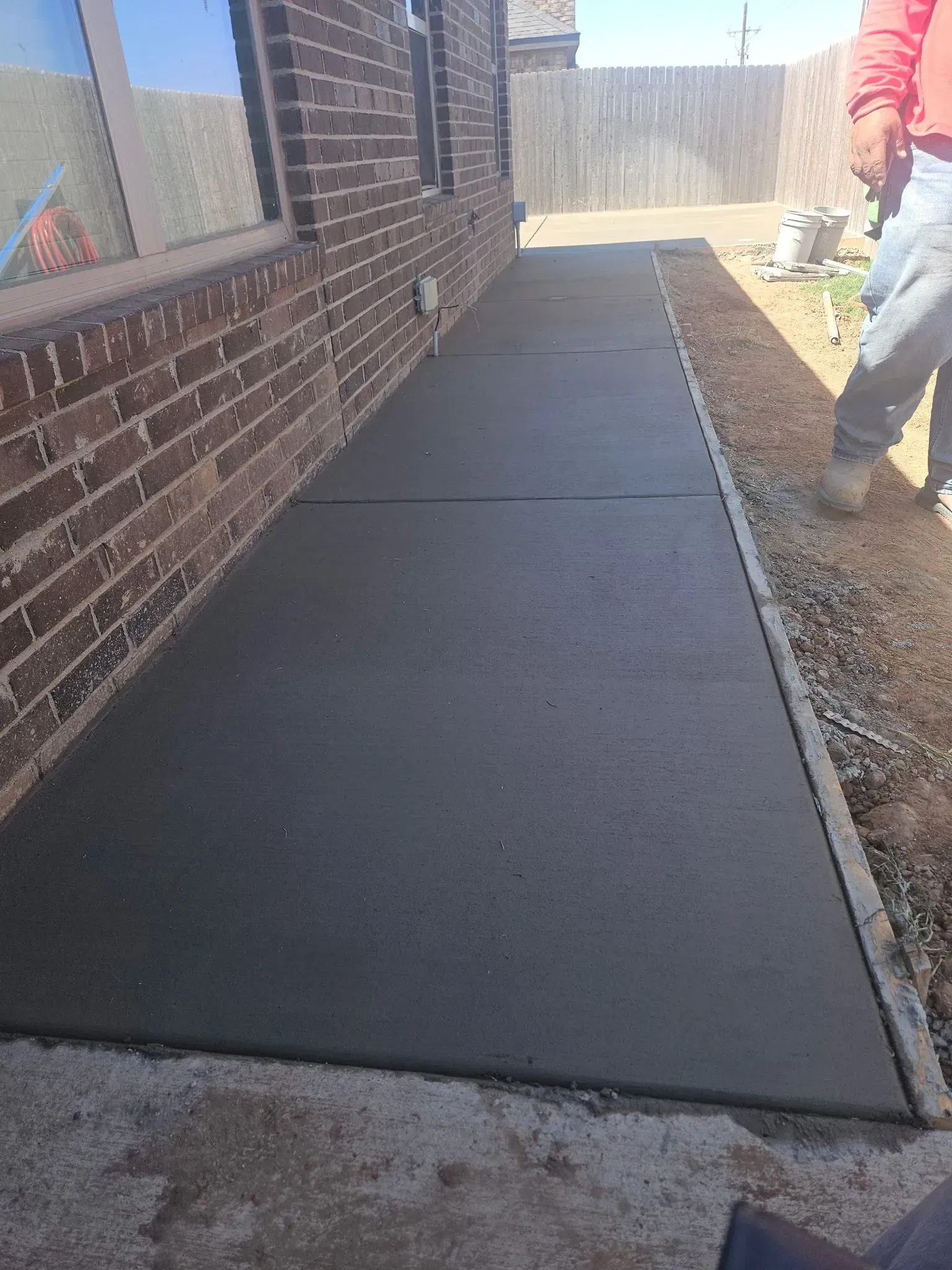 Freshly poured concrete walkway beside a brick house, with a worker at the edge and wooden fence ahead