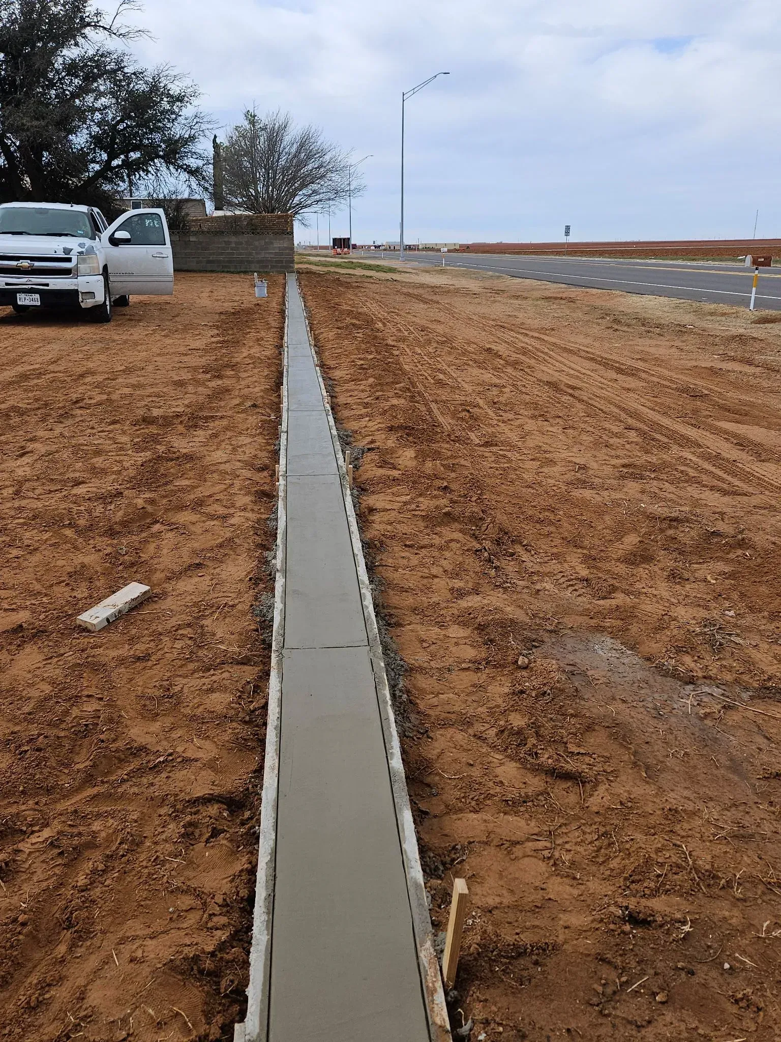 Concrete drainage channel beside a dirt roadside, with parked white trucks and an open road under cloudy sky.