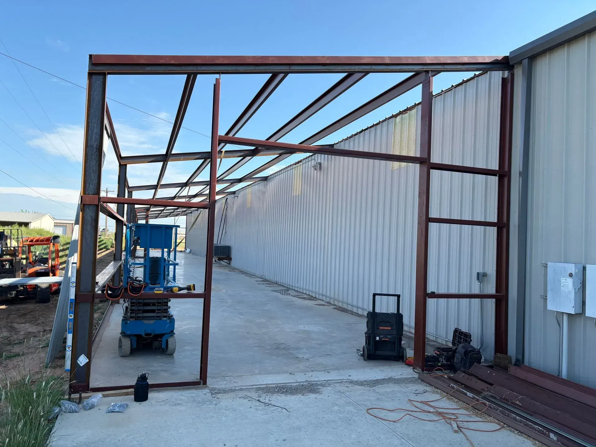 Rusty steel frame beside white container wall in an industrial yard, with a blue cart and equipment underneath