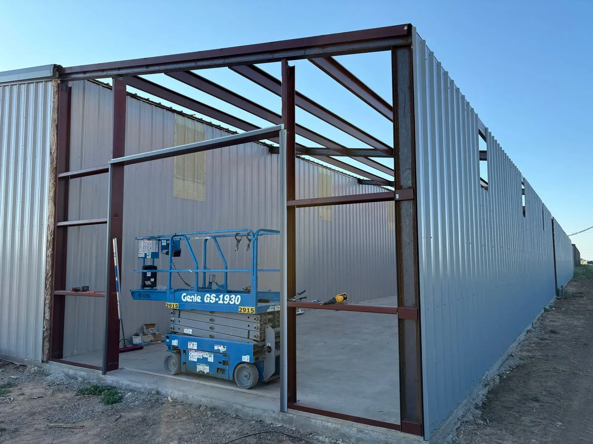 Steel building frame with corrugated metal walls and a blue scissor lift inside under construction.