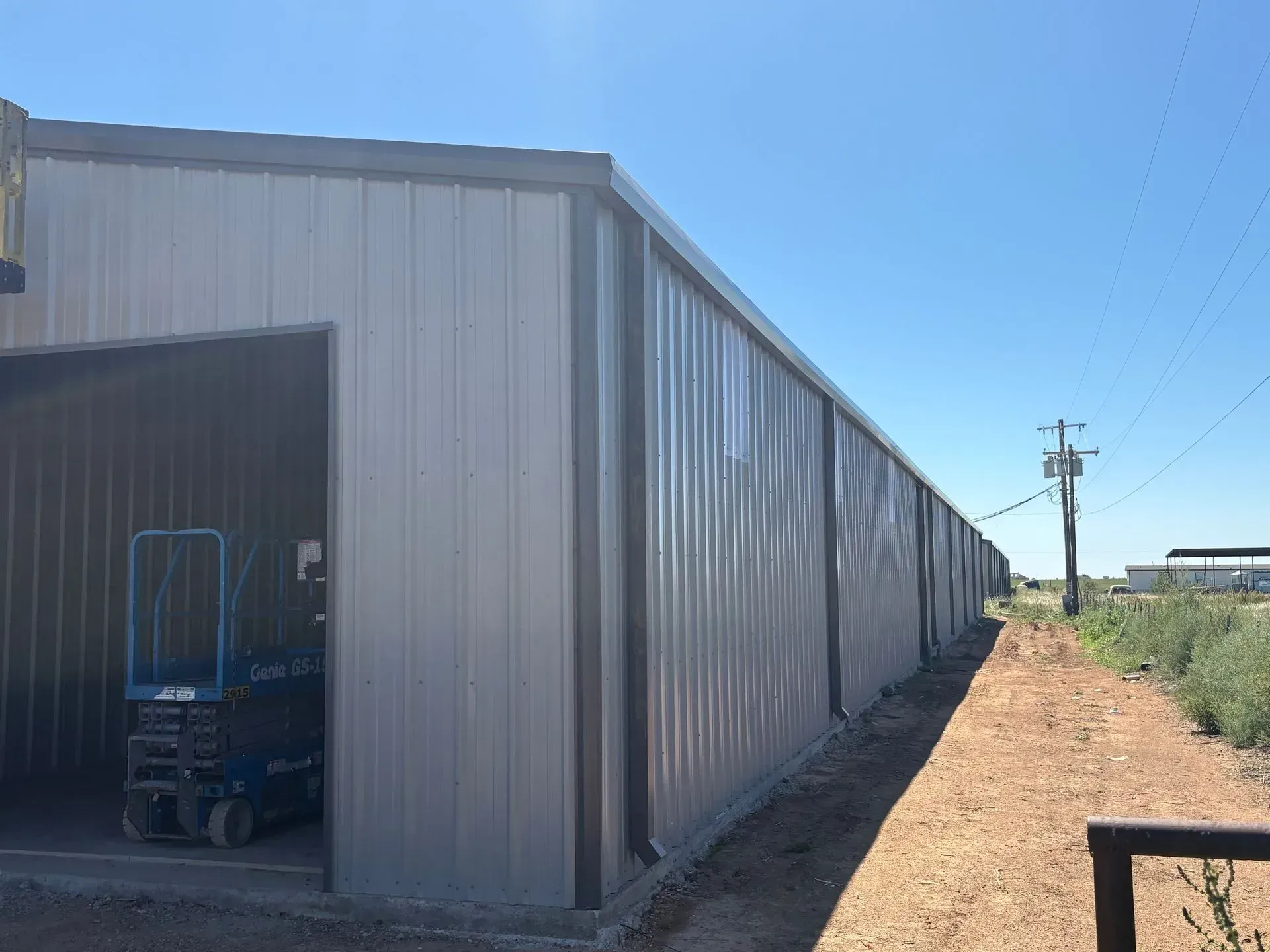 Long metal warehouse with an open door and a blue lift inside, beside a dirt path under a clear sky