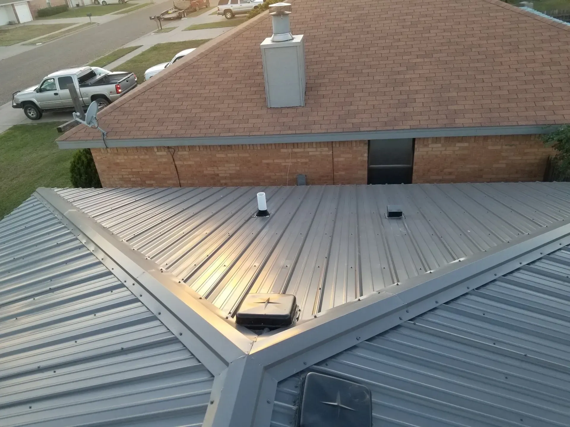 Two adjacent metal roofs meeting at a valley, with a brick house wall and chimney in the background.