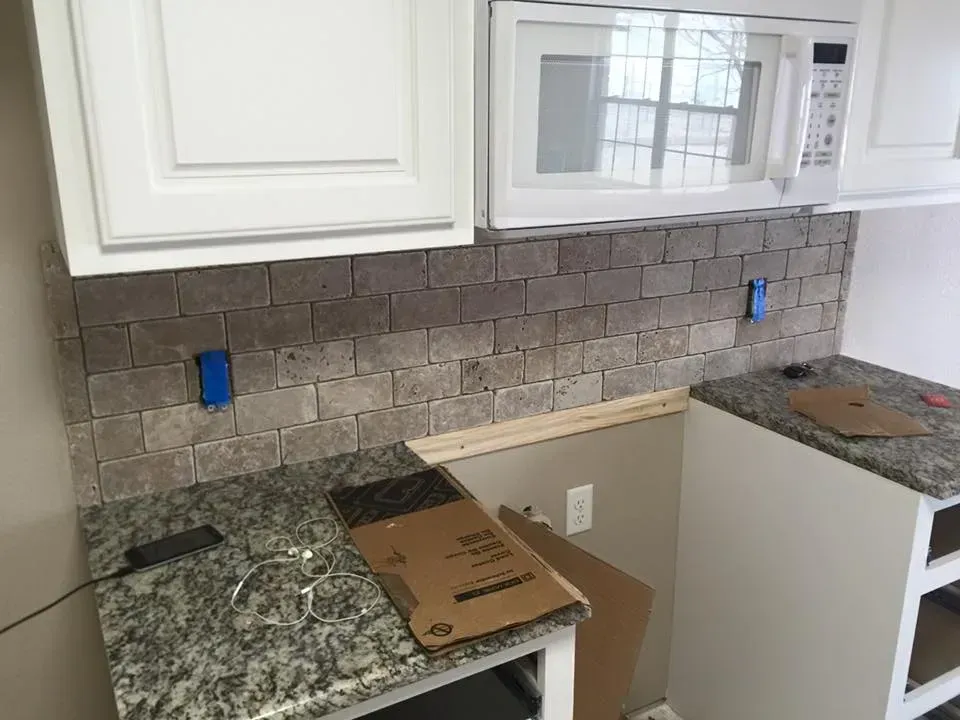Kitchen under renovation with white cabinets, granite counters, tiled backsplash, and cardboard on the counter