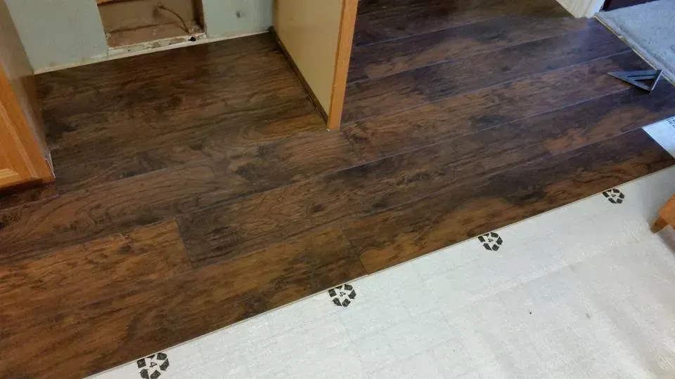Dark wood laminate floor meeting a white tiled surface at a doorway, with an open door visible.